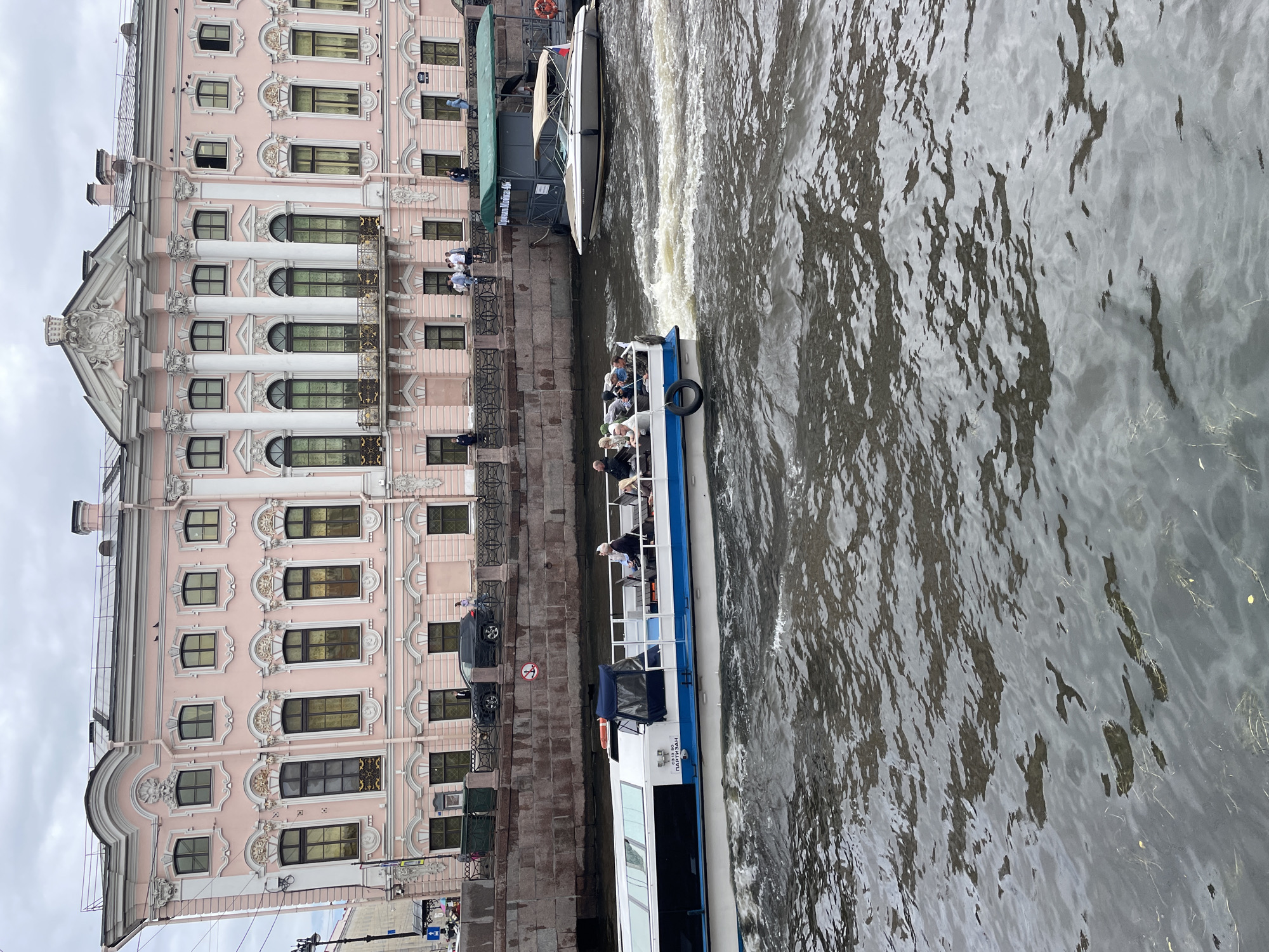 The image depicts a boat tour on a canal in Saint Petersburg, Russia. The boat, labeled "С313 50 ПАРТИЗАН", is carrying several passengers. In the background, a grand pink building with ornate architecture, likely a historic palace or museum, lines the canal. Another smaller boat is docked along the embankment. The canal water is choppy, indicating the movement of the boats. The sky is overcast. The scene captures a typical tourist activity in the city.