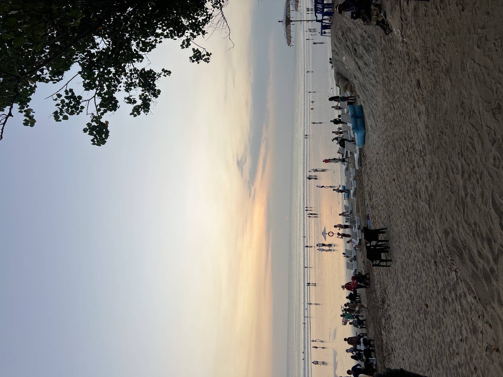 This image depicts a serene beach scene in Kuta, Bali, Indonesia, during the golden hour, just before sunset. The sky is a gradient of soft blues and oranges, with the sun a tiny, distant red disc near the horizon. The ocean is calm, with gentle waves lapping the shore, reflecting the colors of the sky.
The beach is populated by a moderate number of people, engaged in various activities. Some are strolling along the water's edge, others are sitting on lounge chairs or playing in the shallows. In the foreground, a sandy beach is dotted with beach chairs and umbrellas, indicating a popular tourist spot. Several individuals are visible in the foreground, some sitting, others standing.
The overall atmosphere is peaceful and relaxed, characteristic of a popular beach destination at dusk. The presence of lounge chairs, umbrellas, and a lifeguard ring suggests that this is a managed and well-equipped beach area, likely a well-known spot within Kuta. The silhouette of a tree branch with leaves is visible in the upper right corner, adding a natural element to the scene.