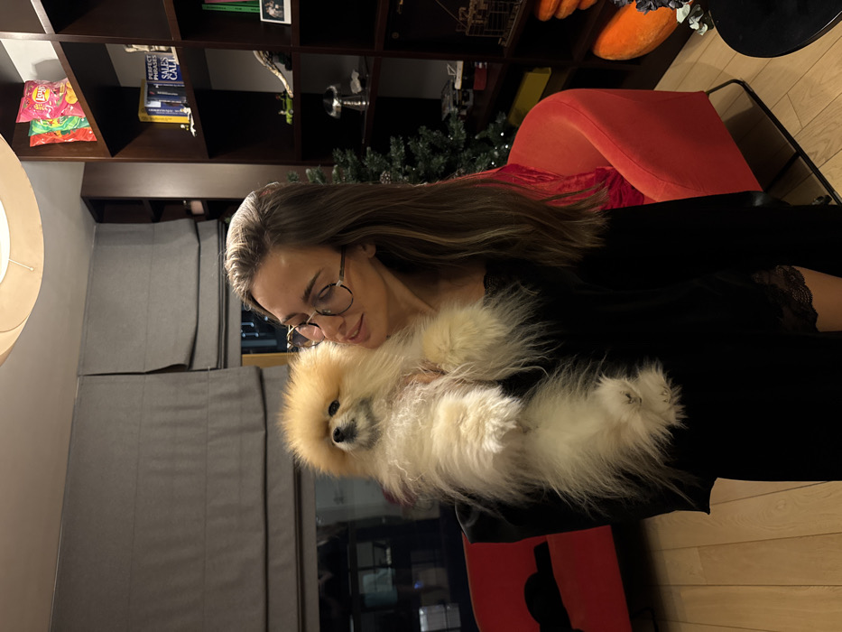 A woman wearing glasses holds a fluffy, light brown Pomeranian dog. She is sitting in an orange chair with dark hair that falls past her shoulders. The background reveals a bookshelf filled with books, decorative objects, and a bag of chips. A gray upholstered wall panel is visible behind her. Hardwood floors are on the right side of the frame.