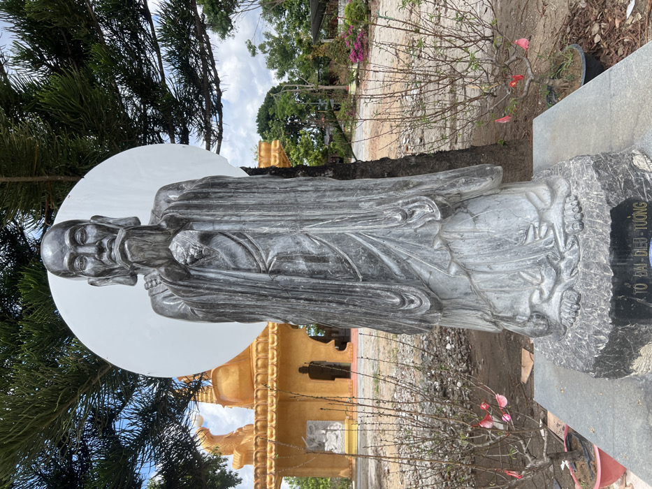 A tall, dark grey stone statue of an elderly male figure with a long beard and flowing robes stands centrally in an outdoor setting in Đăk Mar, Vietnam. The figure's hands are clasped at chest level, and a large, circular, white disc is positioned directly behind its head, resembling a halo. The statue rests on a dark stone base with Vietnamese text inscribed. To the left, a golden multi-tiered architectural structure, likely a temple, is visible, featuring smaller statues within its archways. Sparse, potted deciduous trees with some red/pink blossoms flank the main statue in the foreground. The ground consists of bare earth and patches of green foliage with pink flowering plants in the midground to the right. The background features more trees and utility poles under a partly cloudy sky. No people are visible.