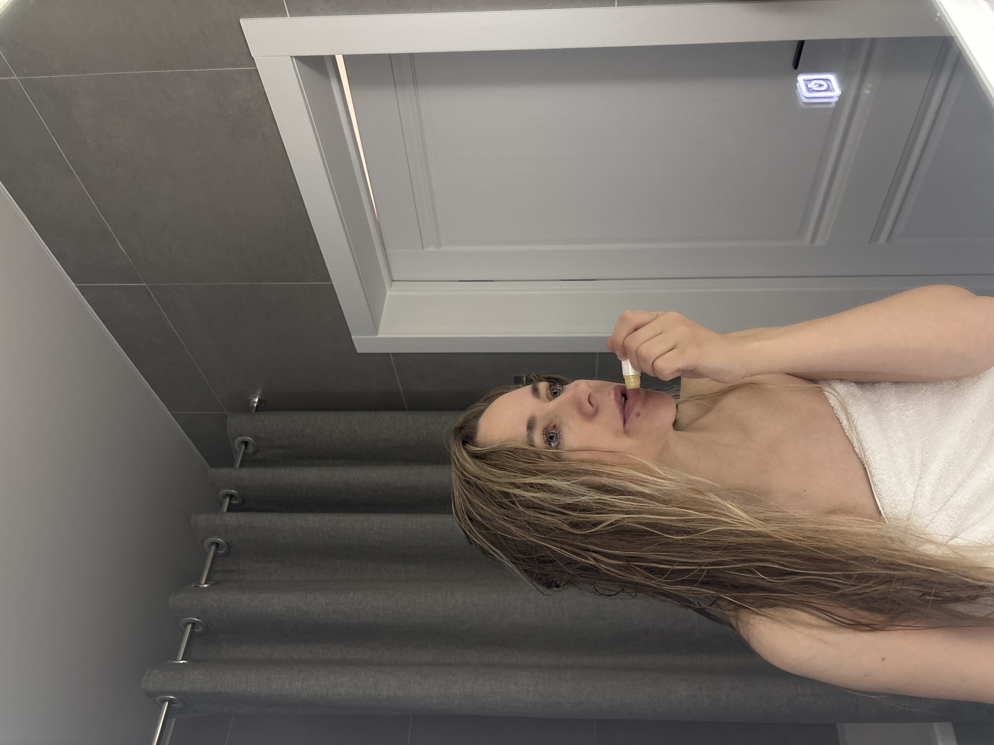 A woman wearing a white towel is applying lip balm while looking in a mirror. The mirror is framed with white trim and has a built-in light. Behind the woman are gray tiled walls and gray pleated curtains. There is no indication of the city of Chaikovsky, Russia.