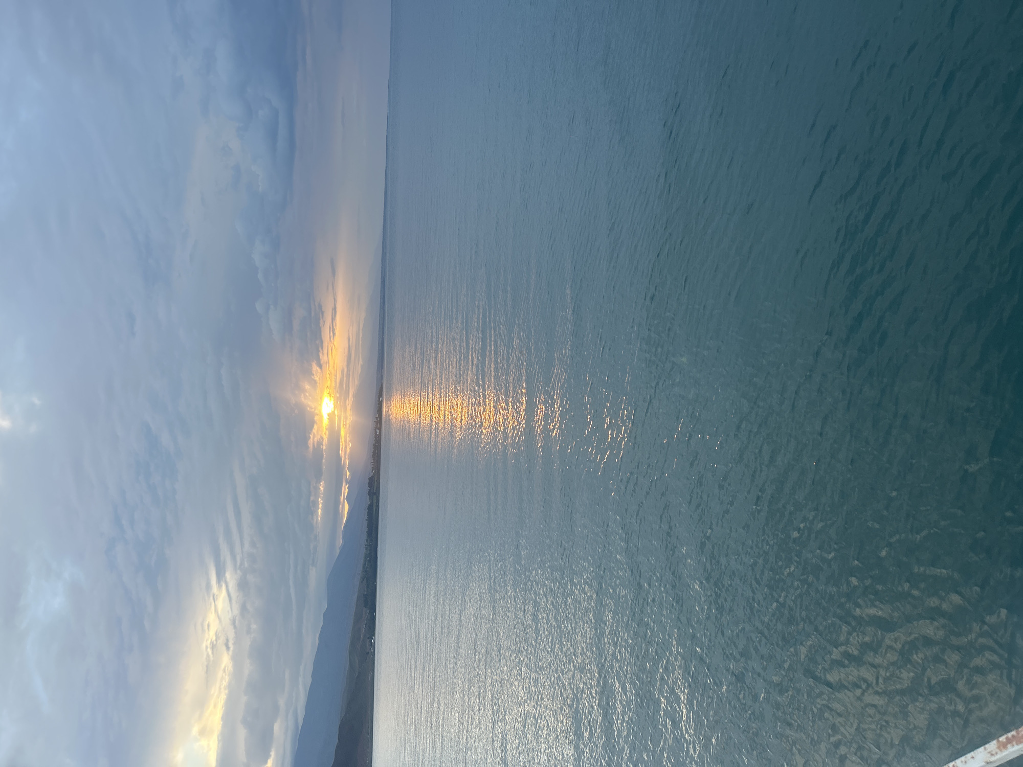The image shows a body of water, likely Issyk-Kul Lake, at sunrise, with the sun breaking through cloudy skies. The water is calm with gentle ripples, reflecting the golden light of the sun. In the distance, a range of mountains lines the horizon. The sky is filled with a mix of gray clouds and patches of lighter, warmer tones where the sun is visible. The foreground shows the edge of a structure, possibly a pier or boat, with a white and rusty metal railing. The overall scene depicts a serene and tranquil natural landscape.