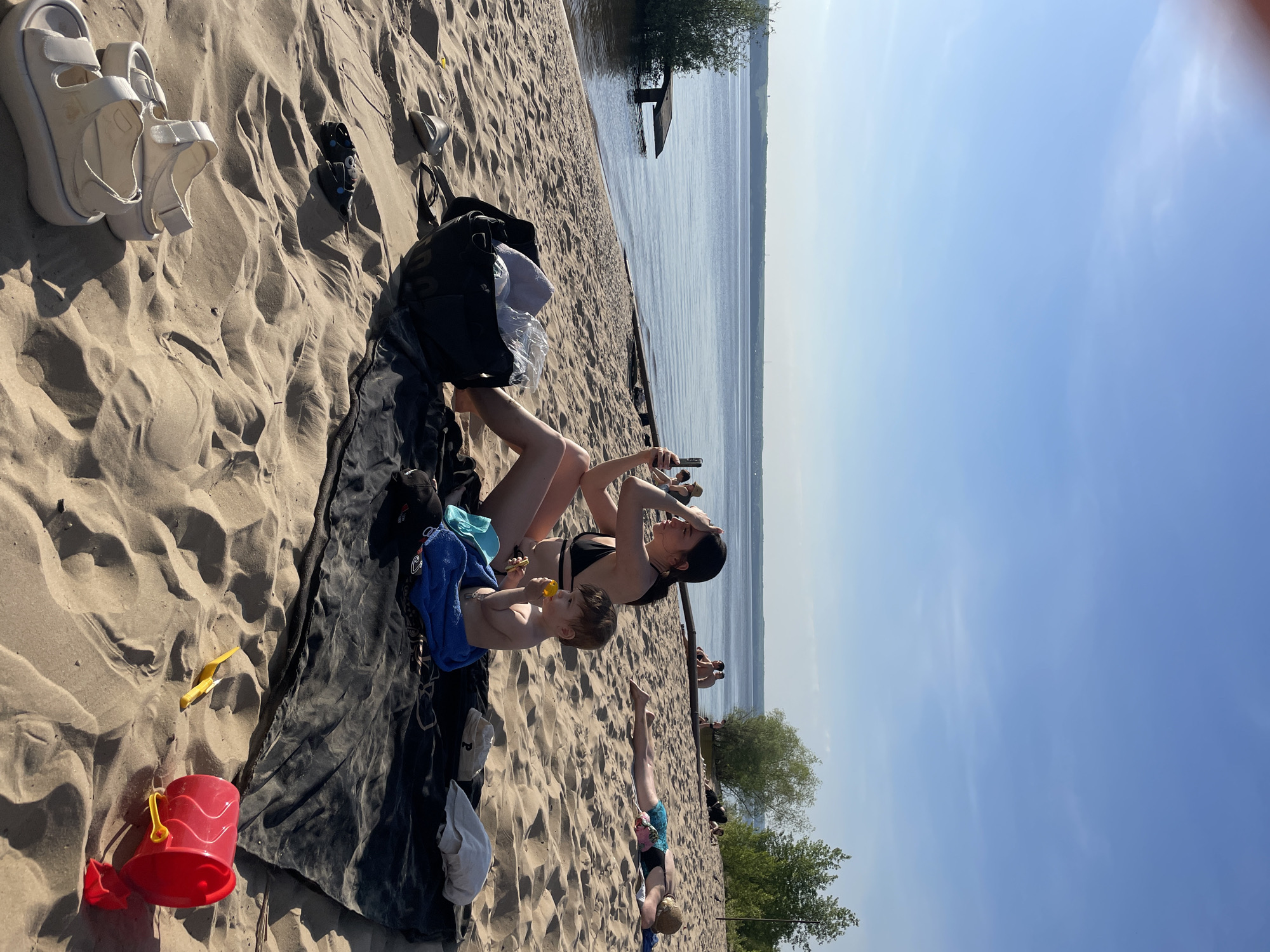 The image depicts a beach scene in Matiushino, Russia. A woman in a black bikini and black cap is seated on a dark blanket with a child. The child is eating and is covered by a blue towel. The woman is holding a mobile phone. Beside them is a black bag, along with white sandals and other footwear in the sand. In the background, there is a body of water, trees, and other people sunbathing. A red bucket and a yellow shovel are visible on the left side of the blanket.