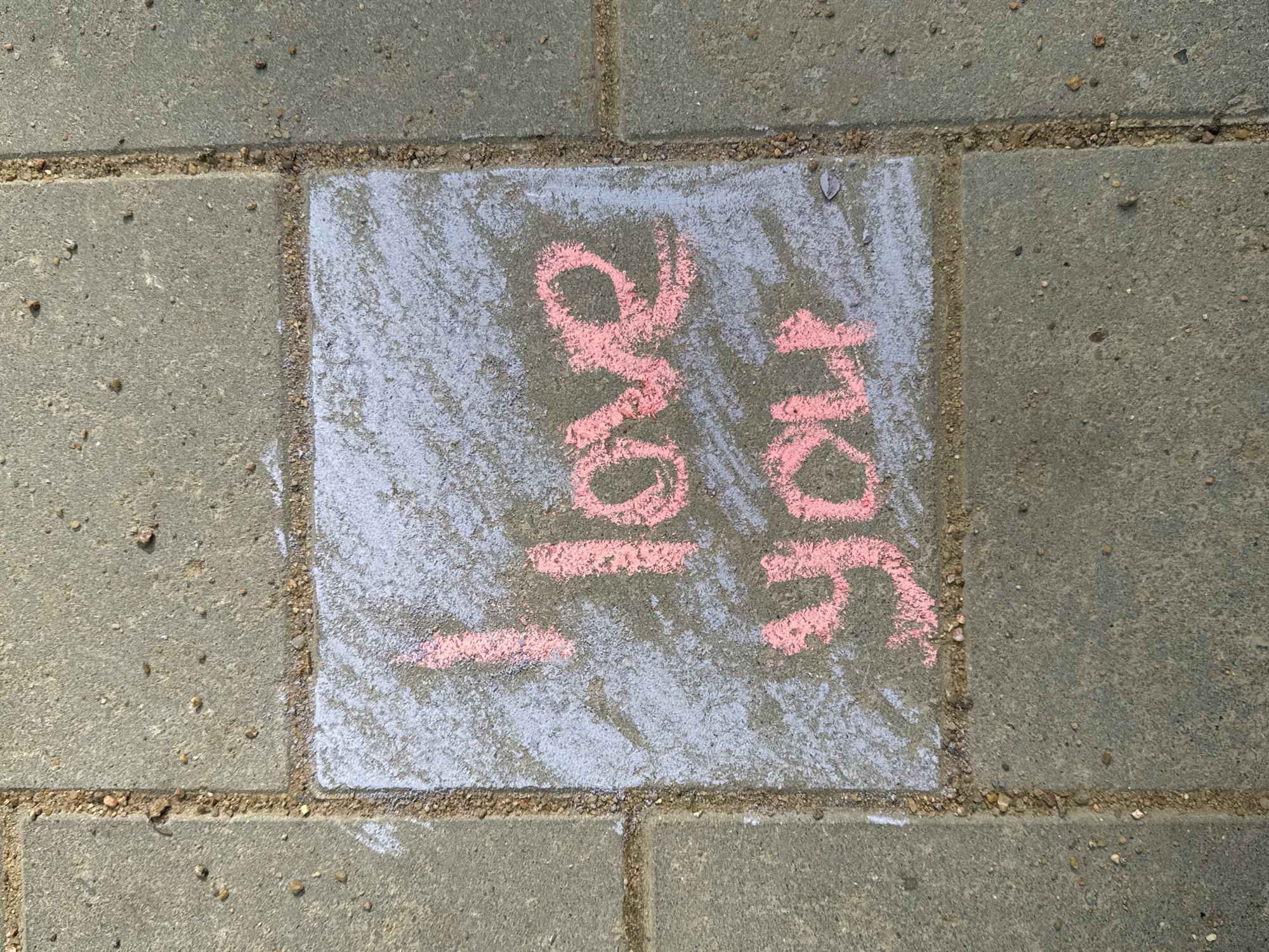 The image shows a light-gray concrete paving stone with pink chalk writing on it. The words "I love you" are written in blocky, stylized letters. The paving stone is surrounded by other similar stones. The area between the stones is filled with dark sand. Based on the light-gray concrete paving stones found throughout the city, and the style of street art, it is possible the image was taken in Saint Petersburg, Russia.