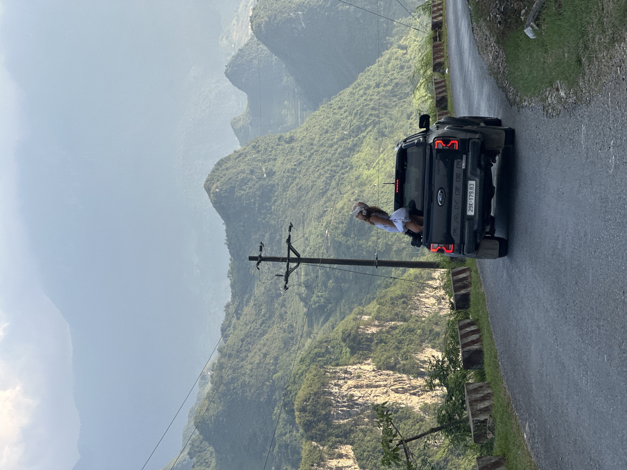 A woman is leaning out of the passenger window of a black Ford Ranger pickup truck that is driving on a winding road. The truck's license plate reads "29K-179.83". In the background are mountains obscured by a hazy sky and an electrical pole. The road is lined with a low concrete barrier. The location is Đường Thượng Commune, Vietnam.