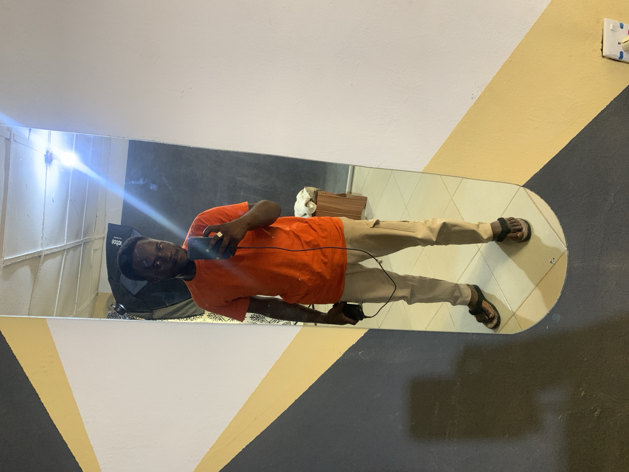A full-body selfie is taken in a mirrored room, likely in Jos, Nigeria, with a striking wall design featuring diagonal bands of black, yellow, and white. The individual, a dark-skinned male, stands centered in the frame, wearing a bright orange t-shirt, khaki cargo pants, and brown sandals. He holds a black smartphone in his right hand, capturing the reflection. A black cord extends from the phone, connecting to an unseen device. In his left hand, he holds a black object, possibly a camera or a flash unit. The background reflected in the mirror reveals studio lighting equipment, a wooden stool with a white towel, and a patterned curtain. The floor appears to be tiled in a cream color. The overall impression is of a personal, possibly creative space.