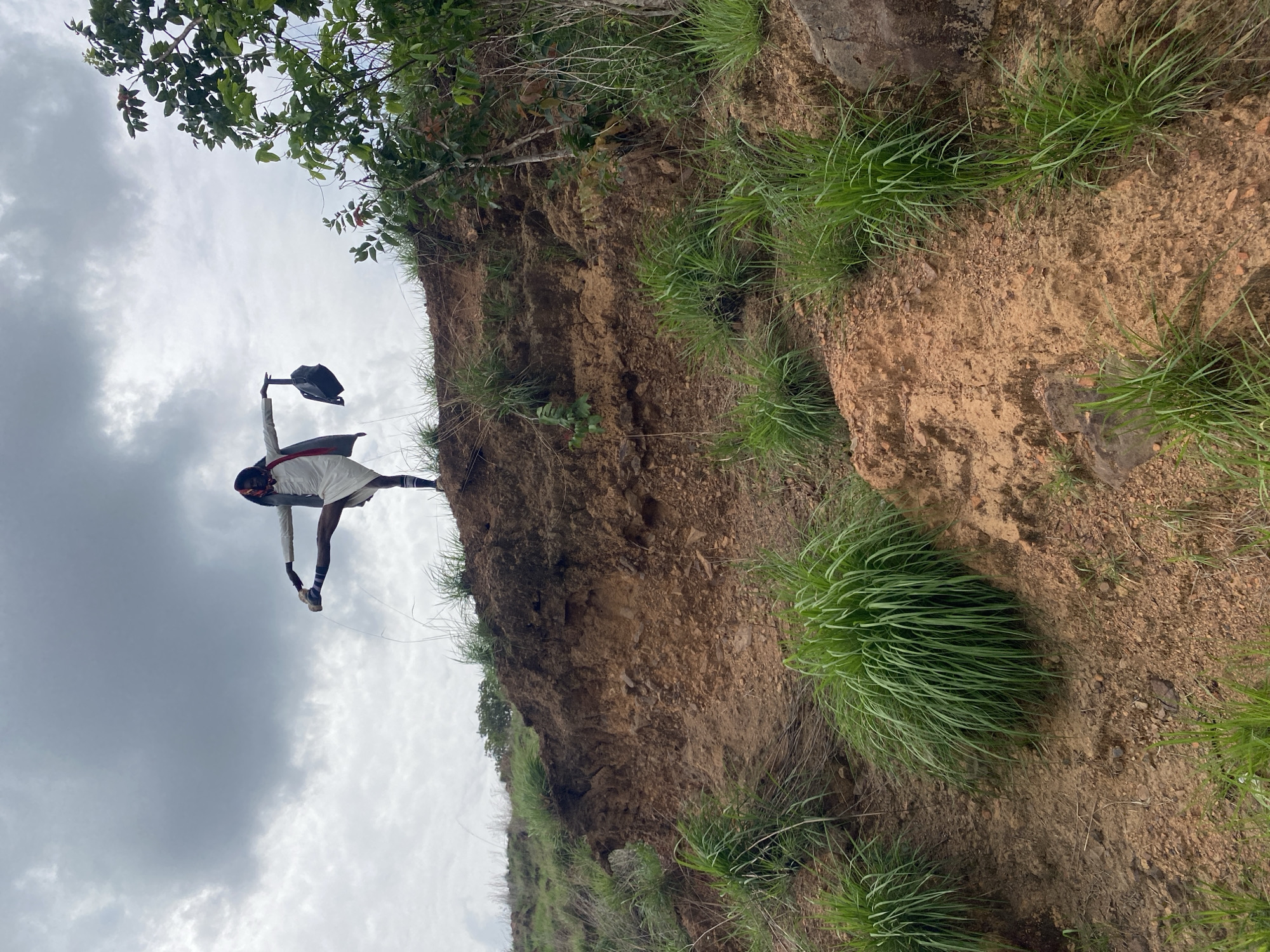 The image, taken from a low angle, shows a person standing on the edge of a dirt cliff or embankment. The person is dark-skinned, wearing a white tunic with black sleeves, socks, and shoes. The person's arms are outstretched and one leg extended, with the person's hand grasping their foot. They also hold a black object, possibly a bag, aloft in one hand.
The cliff is composed of brown earth and sparse green grass, and it rises above the viewer. A tree with green leaves is partially visible in the top left corner of the image. The sky is overcast with gray clouds.
While the prompt mentions Amaigbo, Nigeria, this information is not evident from the visual information provided in the image itself. Therefore, Amaigbo is not a conclusion that can be drawn from the photograph.