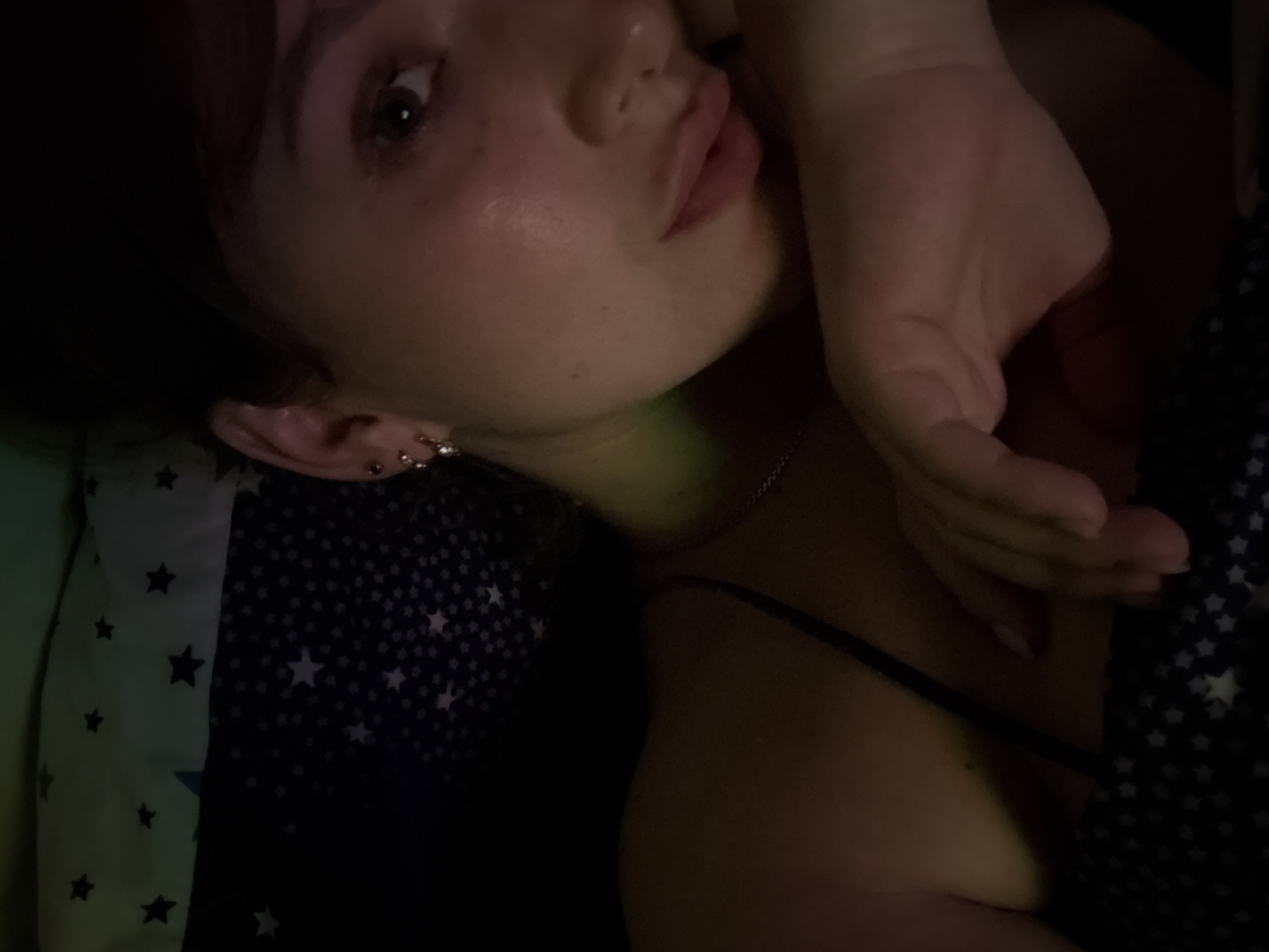 The image is a close-up selfie of a young woman lying down in bed. She is partially obscured by darkness, but her face and shoulder are visible. She has light-colored skin, dark hair, and is wearing earrings. The background appears to be a pillow or blanket with a star pattern. The lighting is dim and appears to be from a screen, casting a greenish glow on her face. The woman is resting her head on her hand, and her eyes are looking towards the camera. The image does not provide any information about the city of Chaikovsky, Russia, or any specific location within it.