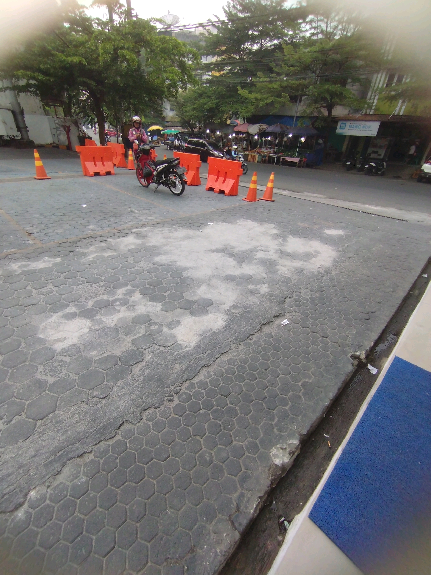 A low-angle, full-shot photograph shows a street scene in Talang Jawa, Indonesia. The foreground is dominated by hexagonal paving stones, some of which are cracked and discolored. To the right, a blue mat and a drainage ditch are visible.
In the mid-ground, a person wearing a pink shirt and a helmet rides a red and black motorcycle. The motorcycle is positioned on the paved street, with several orange traffic barriers and cones scattered around it. Behind the barriers, a market or roadside stalls can be seen with umbrellas and awnings, some with goods displayed on tables. A white banner with the text "MANG MOK" is visible on a building in the background.
The background features lush green trees, buildings, and a cloudy sky. A satellite dish is visible atop one of the buildings. The overall scene suggests a busy urban area with commercial activity.