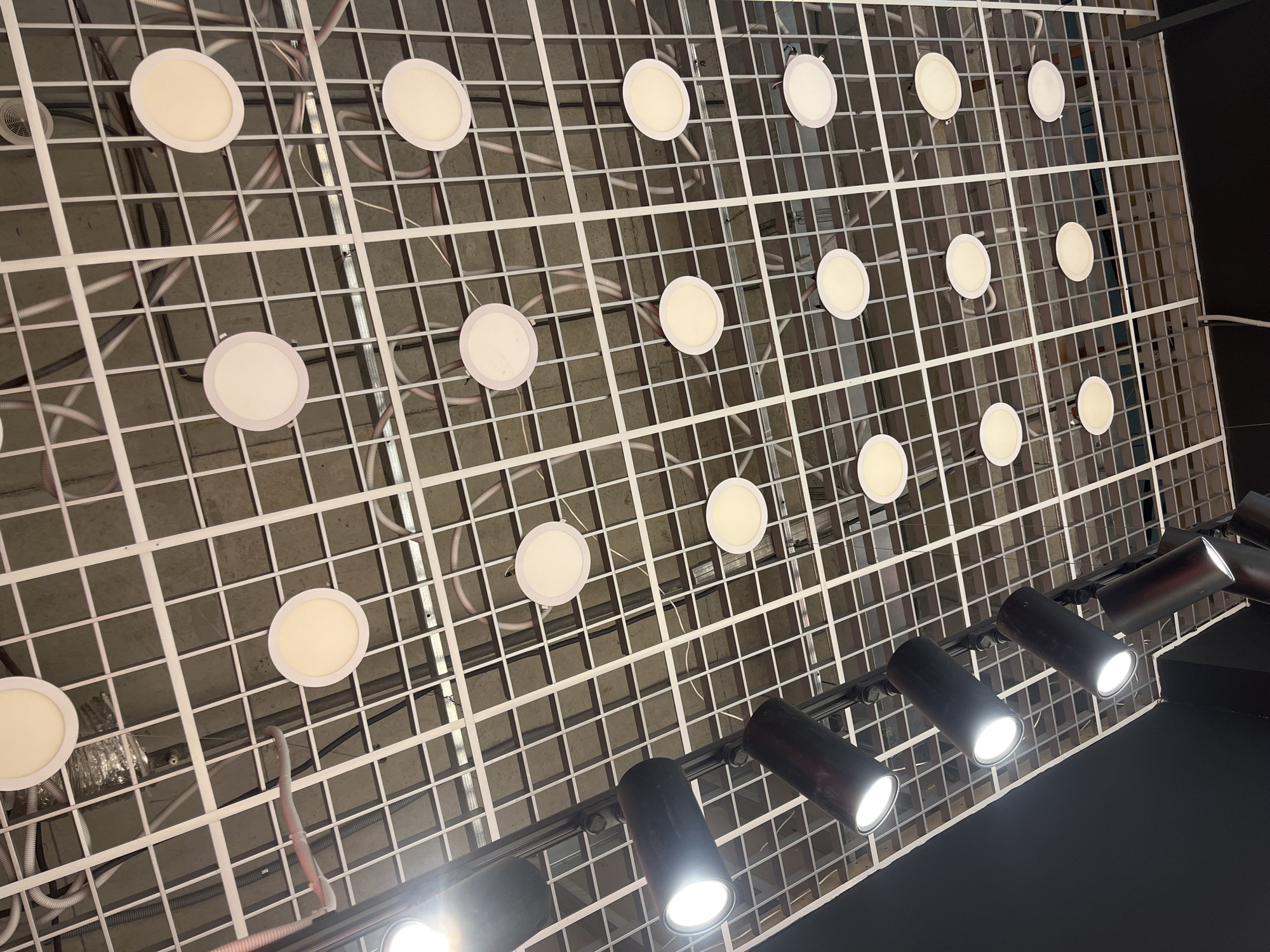 The media file shows a low-angle view of a suspended ceiling with a grid structure. Embedded within the grid are several circular, white LED lights. The lights are evenly spaced across the ceiling, with some appearing to be recessed into the grid. Below the grid ceiling, a black track light system is visible, featuring multiple black cylindrical spotlights, each emitting a bright white light. The spotlights are mounted on the track and are angled downwards. The overall impression is of an indoor space with a modern lighting system, possibly a retail store, exhibition hall, or studio. The lighting suggests it could be daytime or nighttime, as the artificial lights are the primary illumination source visible. There is no visible text in the image.