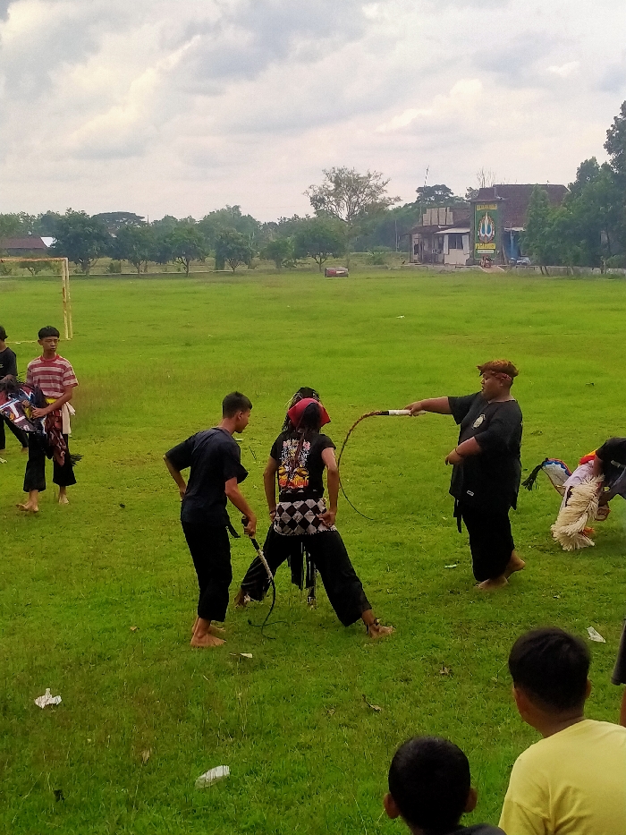 A group of people are on a grassy field, seemingly practicing or performing a traditional activity. Three figures in the center are the main focus, with two holding whips, seemingly engaged in a mock battle. Another individual, dressed in all black and wearing a headdress, appears to be supervising or instructing. Other people stand around, some holding masks or costumes. The backdrop features a building, a soccer goal, and trees. The sky is overcast, suggesting a cloudy day.