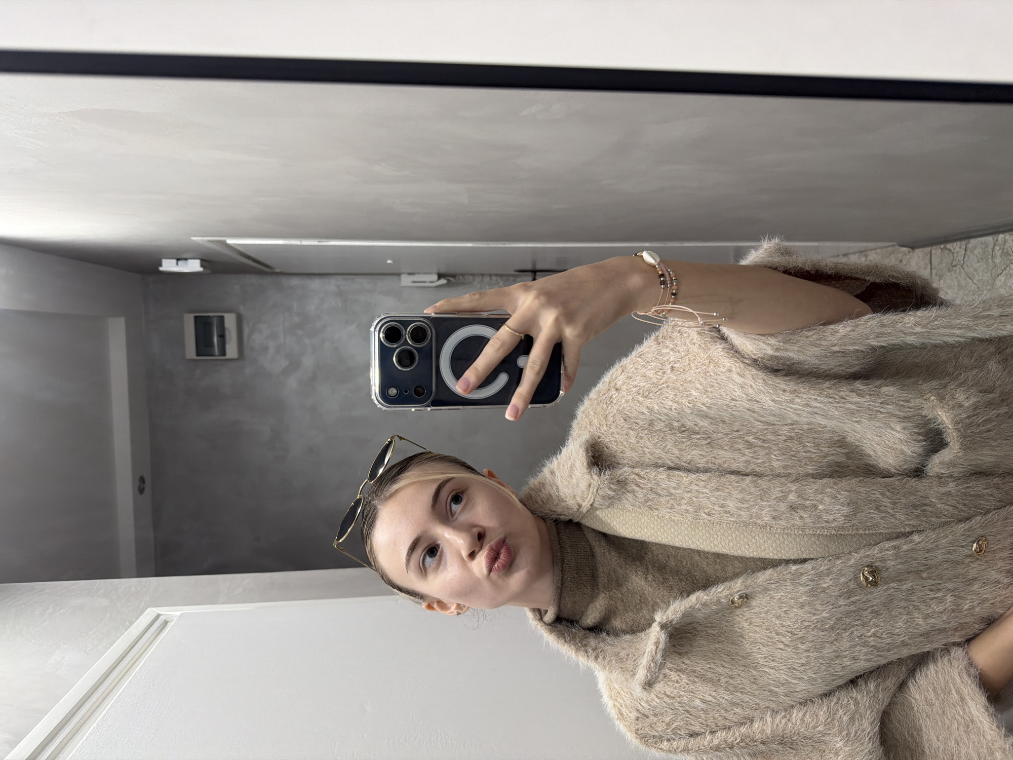 A fair-skinned woman is taking a selfie in a bathroom with a phone featuring a dark case. She's wearing sunglasses perched on her head and a tan sweater with ornate gold buttons. Her hand, adorned with a gold ring and bracelet, holds the phone. The background is a bathroom with a white door and a grey wall featuring a small utility box. The ceiling is white with black trim. The specific location in Plovdiv, Bulgaria cannot be determined from the provided image.