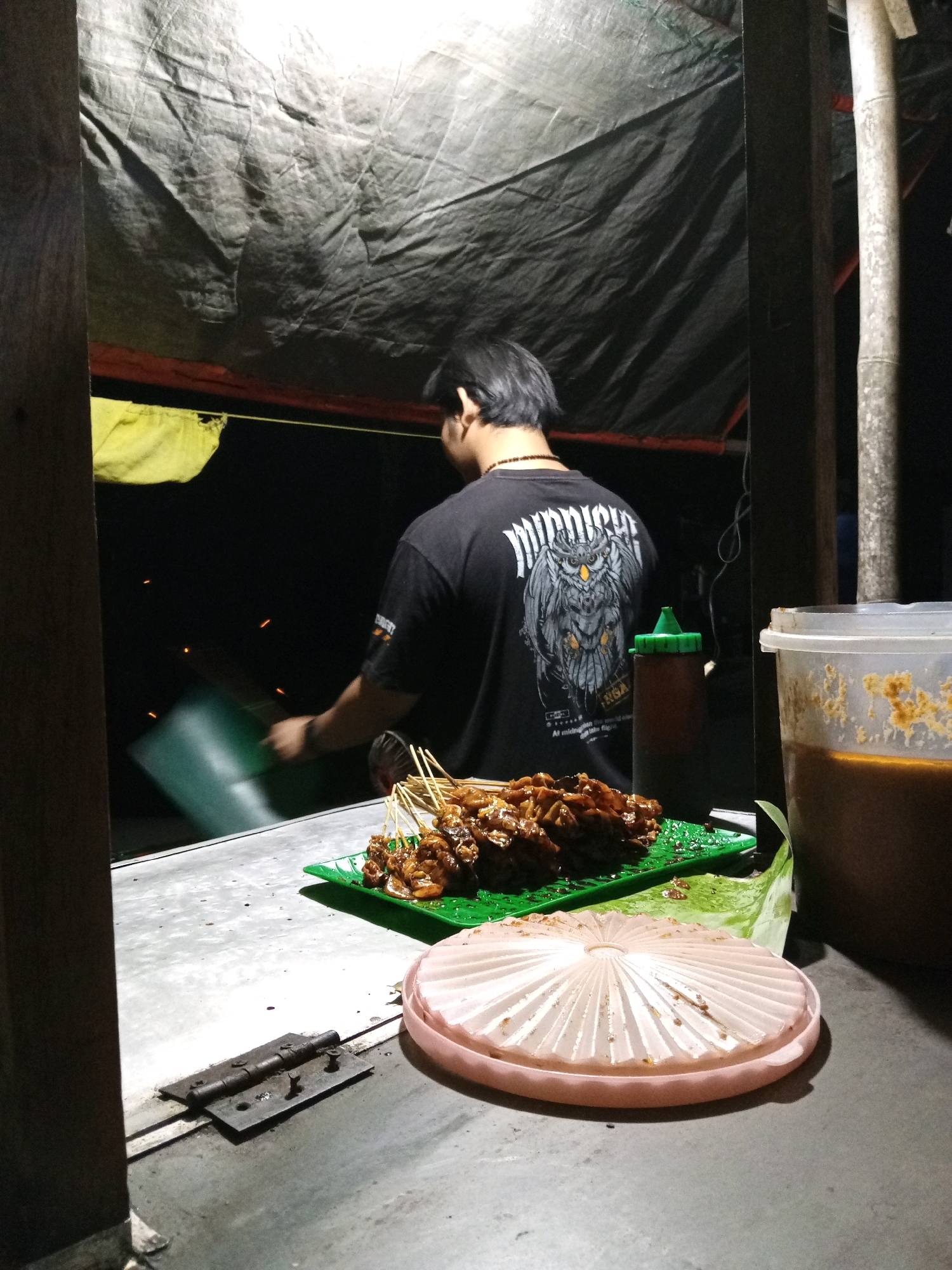 The image shows a street food stall at night in Kedungwinong, Indonesia. A man is standing behind the counter, seen from the back. He is wearing a black t-shirt with a graphic of an owl on it. In front of him, on the counter, is a green plate filled with skewers of grilled meat, likely satay. To the right of the skewers is a container of sauce and a plastic bottle with a green cap. In the foreground, a pink pleated lid rests on the counter. The stall is covered by a dark tarp, and the background is dark, suggesting it is nighttime. The scene depicts a typical outdoor food vendor setup.