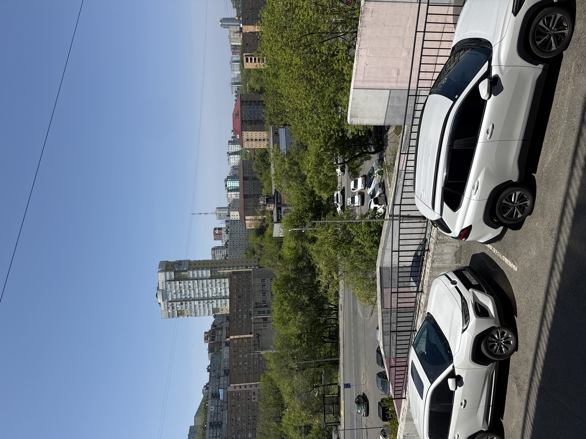 The image shows a view from a high vantage point in Vladivostok, Russia. Two white cars, likely Toyota RAV4 models, are parked closely together on a paved surface, possibly a parking lot or the side of a road. A black metal railing is visible between the viewer and the cars, suggesting the perspective is from a balcony or similar elevated position. Beyond the parked cars, the scene reveals a cityscape with numerous apartment buildings of varying heights and architectural styles. Lush green trees fill the spaces between the buildings. A road runs through the cityscape, with a few other vehicles visible in the distance. The sky is a clear, bright blue. The scene is well-lit, indicating it is daytime.