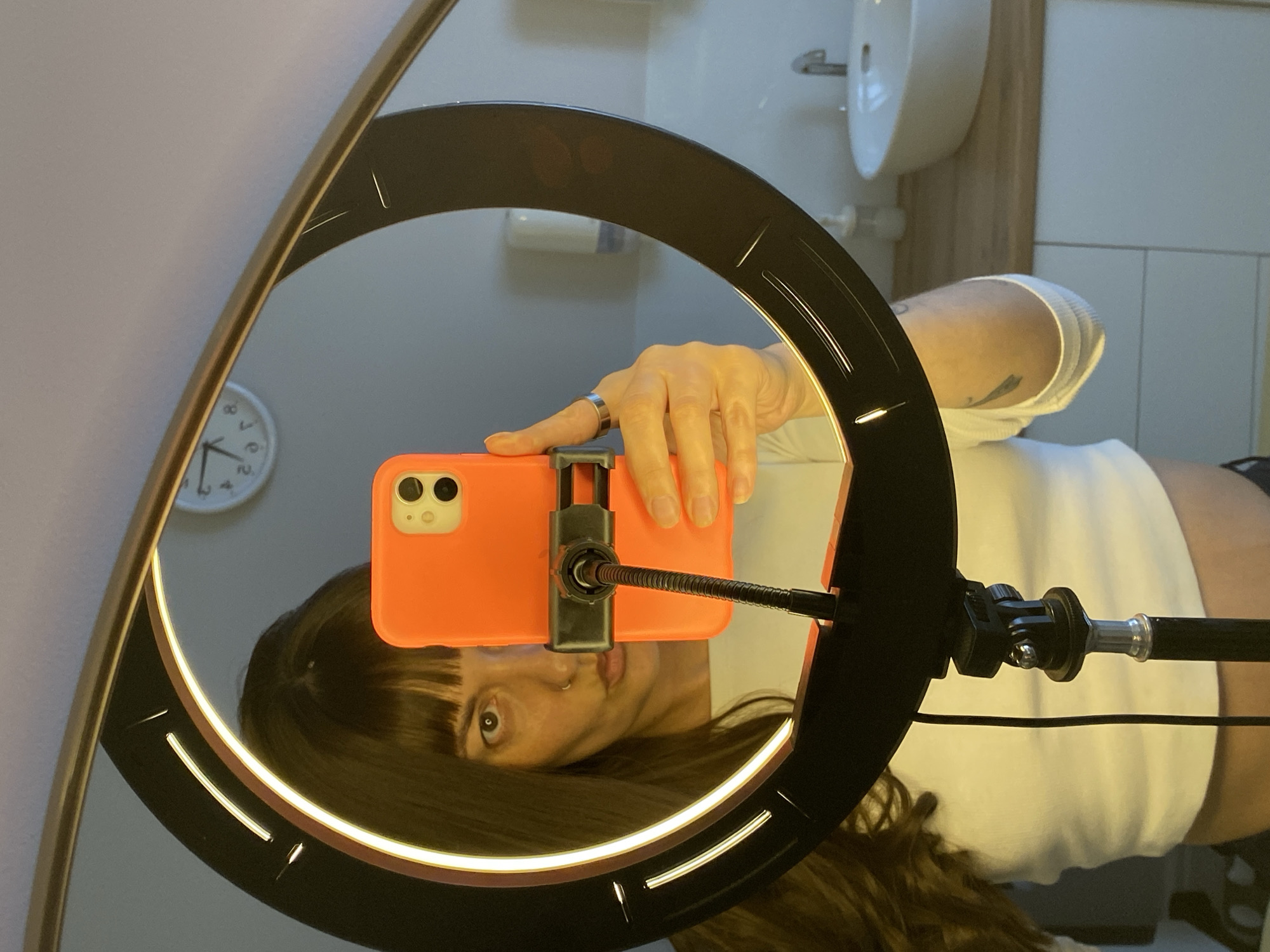 A woman is taking a selfie in a mirror using a phone attached to a ring light. The reflection reveals a bathroom setting with a sink, tiled walls, and a clock. The woman is holding the phone with her right hand. The environment seems to be indoors, possibly a residential bathroom. There is no verifiable information to confirm that this photo was taken in Votkinsk, Russia.