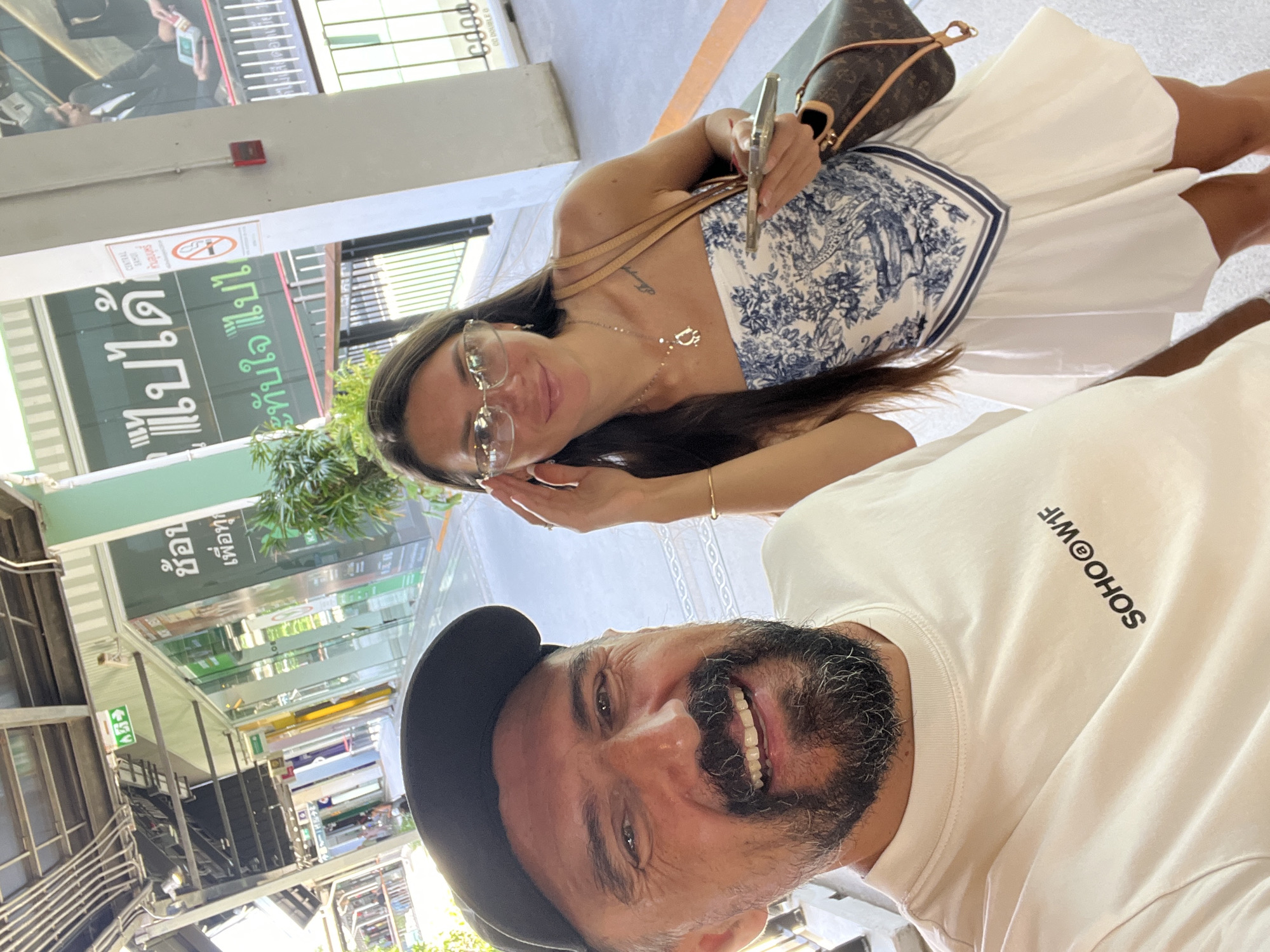 This is a selfie taken by a man and a woman in an outdoor, urban environment. The man, wearing a black baseball cap and a white t-shirt with "SOHO@W1F" printed on it, is in the foreground, smiling and looking at the camera. The woman, wearing glasses and a blue and white patterned strapless top with a white skirt, is positioned behind and to the right of the man. She is adjusting her glasses with one hand and holding a smartphone in the other. A brown designer handbag is slung over her shoulder.
The background suggests a commercial area, possibly a shopping mall or a street with shops. Signs with Thai text are visible on a building, one of which appears to be a "no smoking" sign. The architecture includes exposed beams and pipes overhead, indicating an open-air or partially covered walkway. The ground appears to be paved. Based on the Thai text and architecture, this location is likely in a city in Thailand, potentially in the area of Ban Bang Rak, known for its shopping and commercial establishments. The lighting indicates daytime.