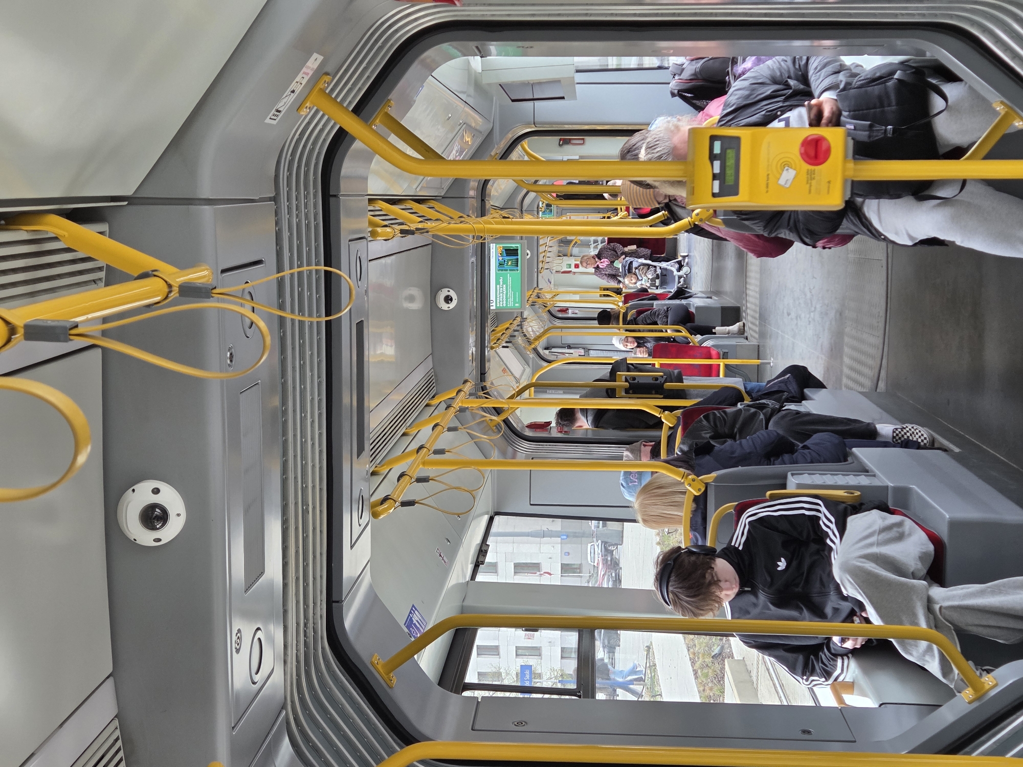 The image shows the interior of a tram or light rail car, likely in motion. Passengers are seated along the sides of the aisle. Yellow handrails and poles run the length of the car. Windows on the right side provide a view of buildings outside, possibly in a city. Based on the "pl. Słowa Sady" sign visible outside the window, it can be deduced that the location is Warsaw, Poland, since "pl." is an abbreviation of "plac" in Polish, meaning square. The color scheme of the tram is gray and yellow. There are surveillance cameras mounted on the interior walls of the tram. Some passengers are looking at their phones, and one is pressing a device with a red button.