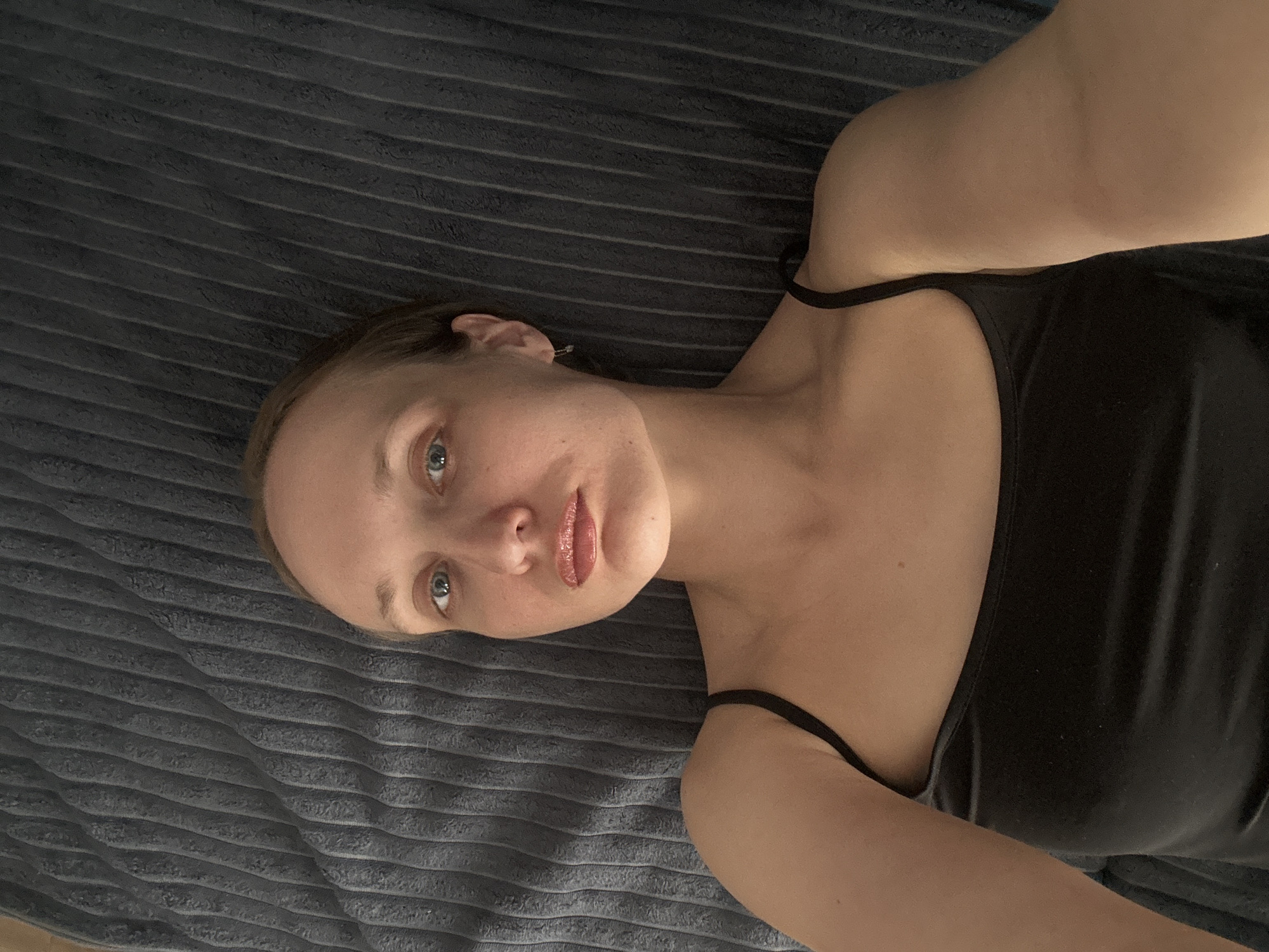 A woman is lying on a grey, horizontally striped blanket. She is wearing a black tank top. Her head is turned towards the camera, her eyes are open, and she is wearing lip gloss. No evidence suggests the location is Подшивалово, Russia.