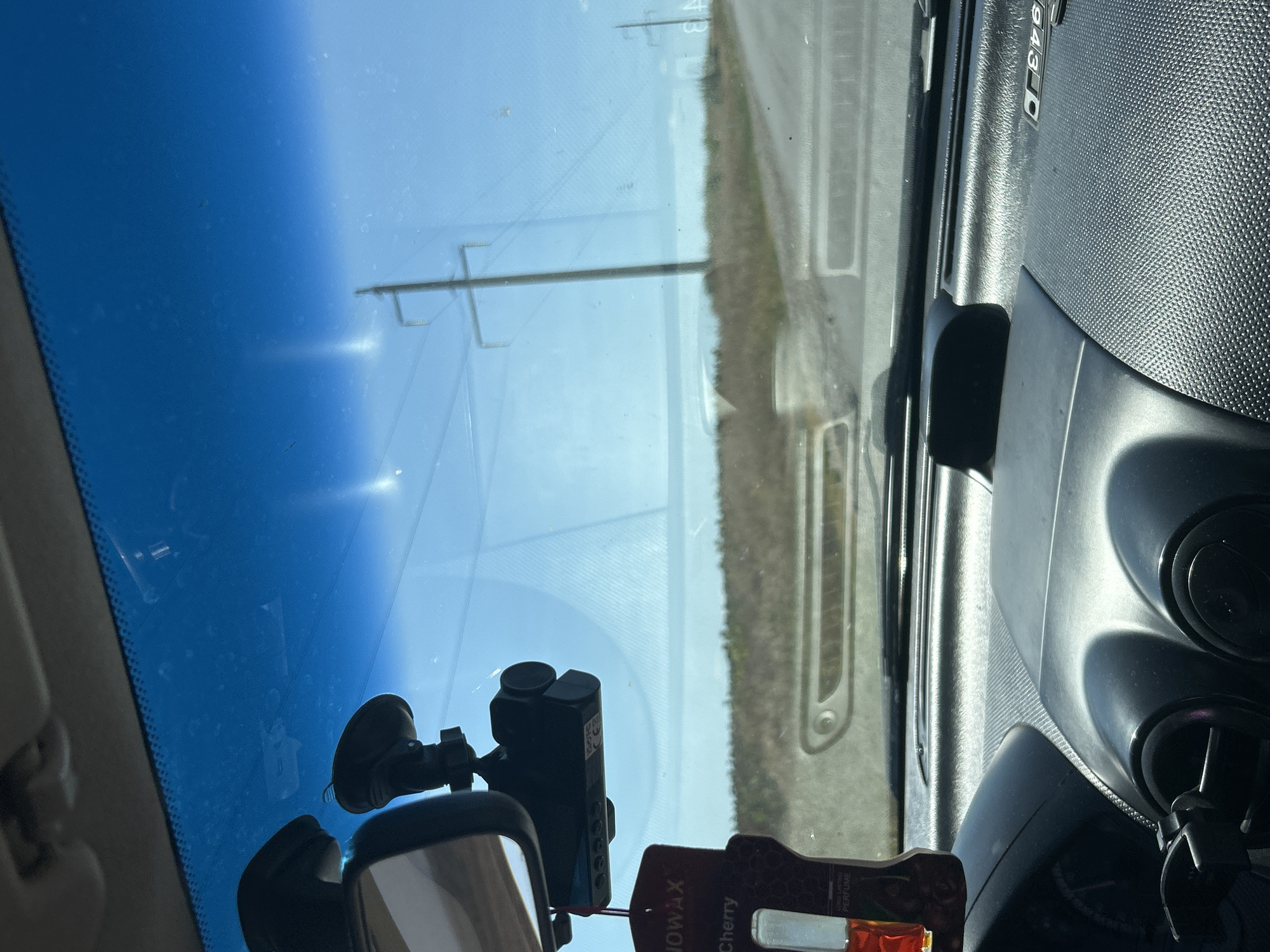 The image shows the interior of a car looking out through the windshield. Mounted on the windshield is a dashcam, and a rear-view mirror is visible to the left. A car air freshener hangs from the mirror. Outside the car, the landscape consists of dry grass and a road, with telephone poles in the distance. The sky is clear and blue. The scene appears to be in a rural or semi-rural area, consistent with the outskirts of Comrat, Moldova.