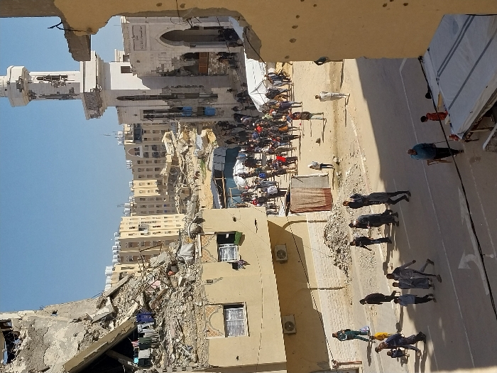This image depicts a severely damaged urban environment in Khan Yunis, Palestinian Territory, under a clear blue sky. The scene is observed from an elevated position behind a damaged, yellow-painted wall with multiple visible perforations.
In the left foreground, a multi-story concrete building is partially collapsed, exposing rebar and internal structures; clothing is hanging to dry from a damaged section. Below it, a two-story building with air conditioning units and barred windows shows less overt damage. A street runs across the midground, marked with a white arrow, where approximately ten individuals, including adults and children, are walking. A man in a green tracksuit stands near a small pile of rubble on the sidewalk. Another man uses a cane while walking in the foreground.
The central midground features a large gathering of people in an open area filled with debris and several white tents. Beyond this crowd, numerous multi-story buildings, many appearing significantly damaged or reduced to rubble, extend into the distance.
In the right midground, a white mosque with a tall minaret stands prominently. Both the mosque's facade and its minaret exhibit extensive damage, including large sections of missing material and structural compromise. Arabic text is visible on the mosque's exterior.
The distant background consists of numerous light-colored residential or commercial buildings, some appearing relatively intact while others show signs of damage.