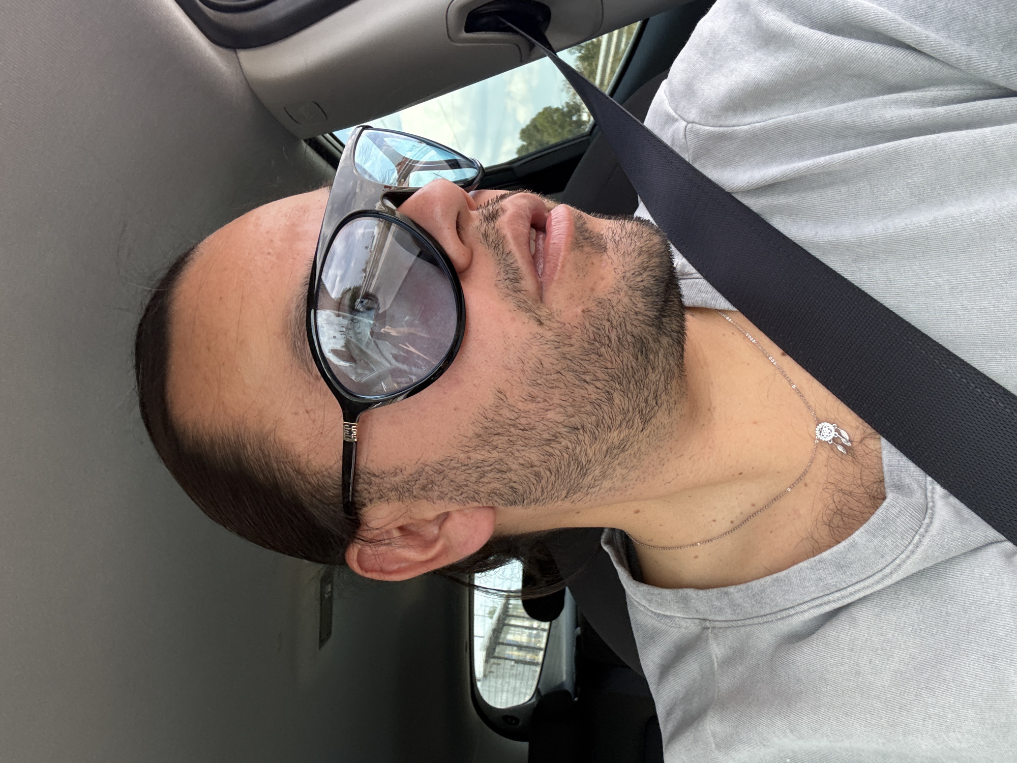 A man with tan skin, facial hair, and a partially bald head is seen in a car, wearing oversized sunglasses and a grey t-shirt. He is wearing a black seatbelt and a silver necklace with a dreamcatcher pendant. The reflection in his sunglasses suggests a building and sky. The interior of the car is visible, with grey panels. Based on the location details, this scene could be inside a car in the Barrio de Nuevo León area of Mexico.