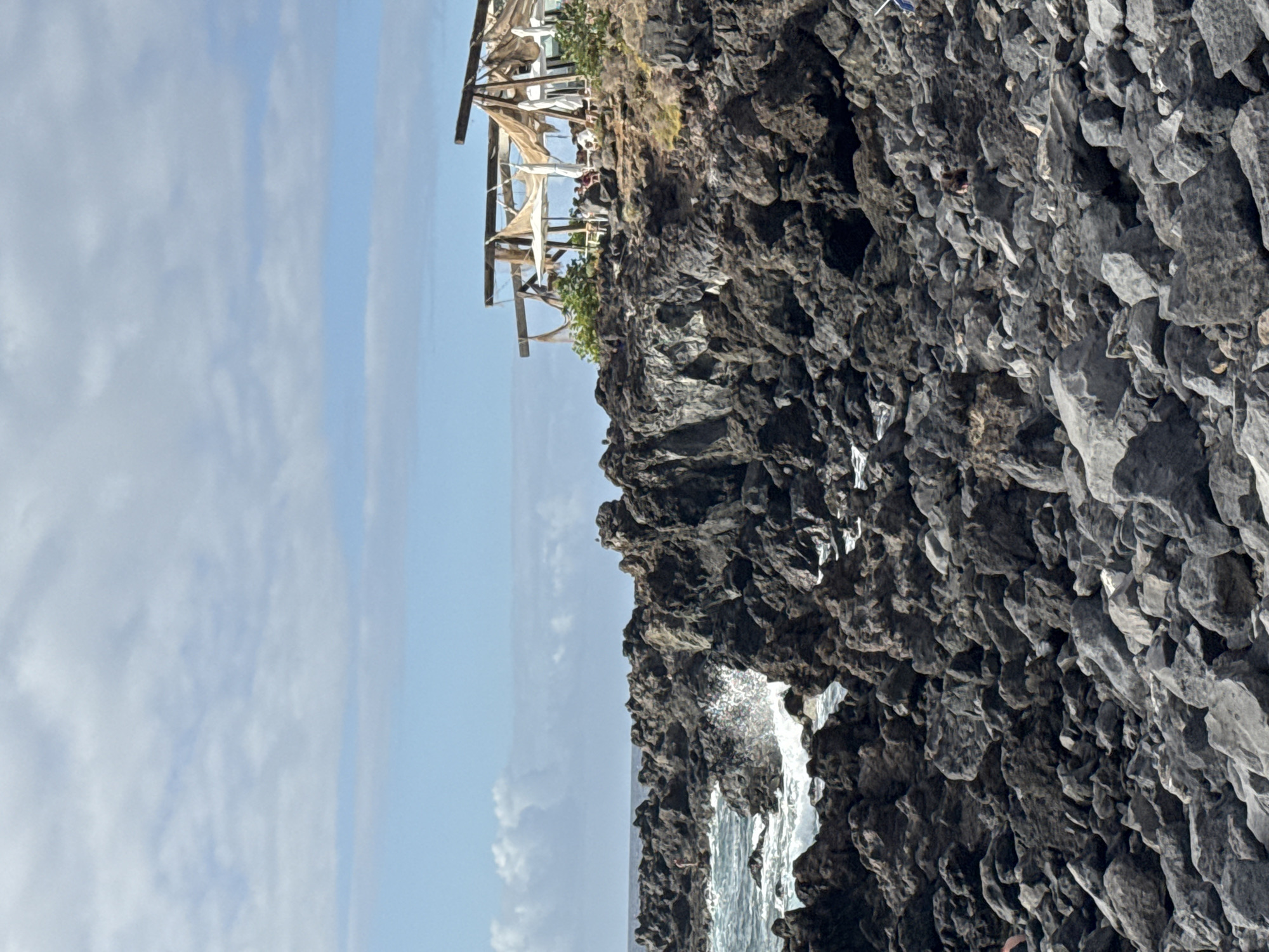 A rocky coastline with a restaurant situated on a cliff overlooking the ocean. The restaurant is open-air, with thatched roofs and patrons seated at tables. Waves crash against the rocks below. The sky is blue with scattered clouds.