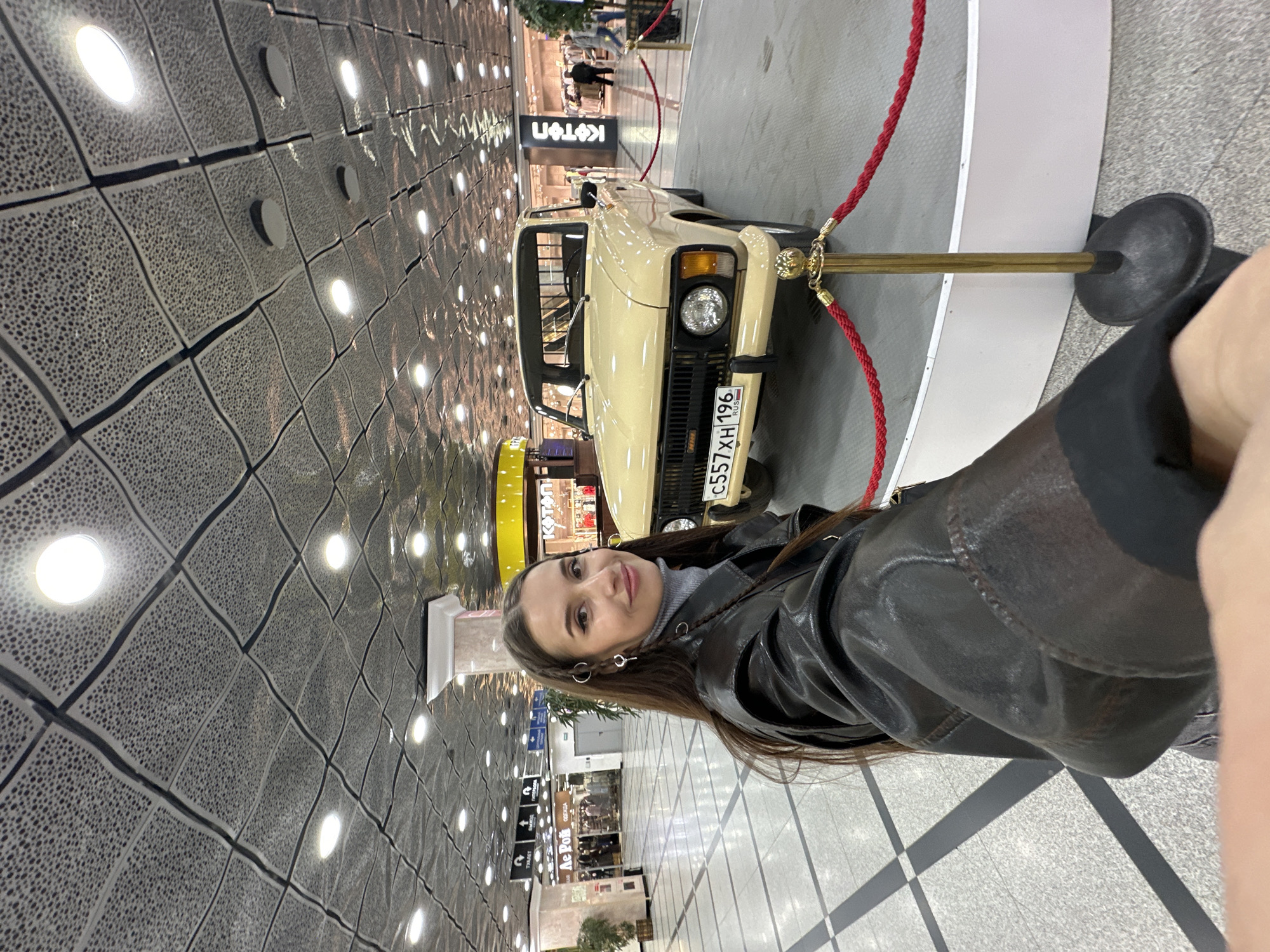 The image captures a selfie of a young woman inside a modern public space, likely a shopping mall in Yekaterinburg, Russia. The woman, with long brown hair, is in the foreground, smiling gently at the camera. She wears a dark brown or black leather jacket over a grey top, and decorative silver rings can be seen in her hair near her left ear, along with earrings. Her arm holding the camera is visible on the far right.
Behind her, a cream-colored vintage or classic car is displayed on a raised platform, cordoned off by red velvet ropes and golden stanchions. The car's front is clearly visible, featuring its grille, headlights, and a Russian license plate that reads "C557XH 196 RUS". The "196" region code corresponds to Sverdlovsk Oblast, where Yekaterinburg is located.
The ceiling above is a notable detail, featuring a unique textured, undulating square pattern with numerous embedded circular lights that reflect off its surface. The floor is tiled with a light-colored, possibly checkered design. In the blurred background, other people are visible, indicating a busy environment. Store signs written in Cyrillic are also discernible, including "KOTON" (a clothing brand) and "ЛЕ РУА" (Le Roy, likely a department store or similar establishment), reinforcing the setting as a commercial building. The lighting suggests it is daytime indoors.