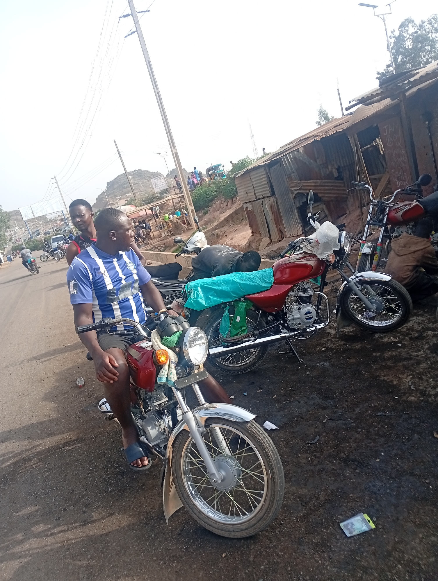 The image depicts two men on a red motorcycle on a road in Jos, Nigeria. The man in the front is wearing a blue and white striped shirt, and the other is wearing a red top. Two other motorcycles are parked nearby. The background shows a corrugated iron structure and a hill in the distance. The location appears to be a street with other people visible in the background. There is trash on the ground near the motorcycles.