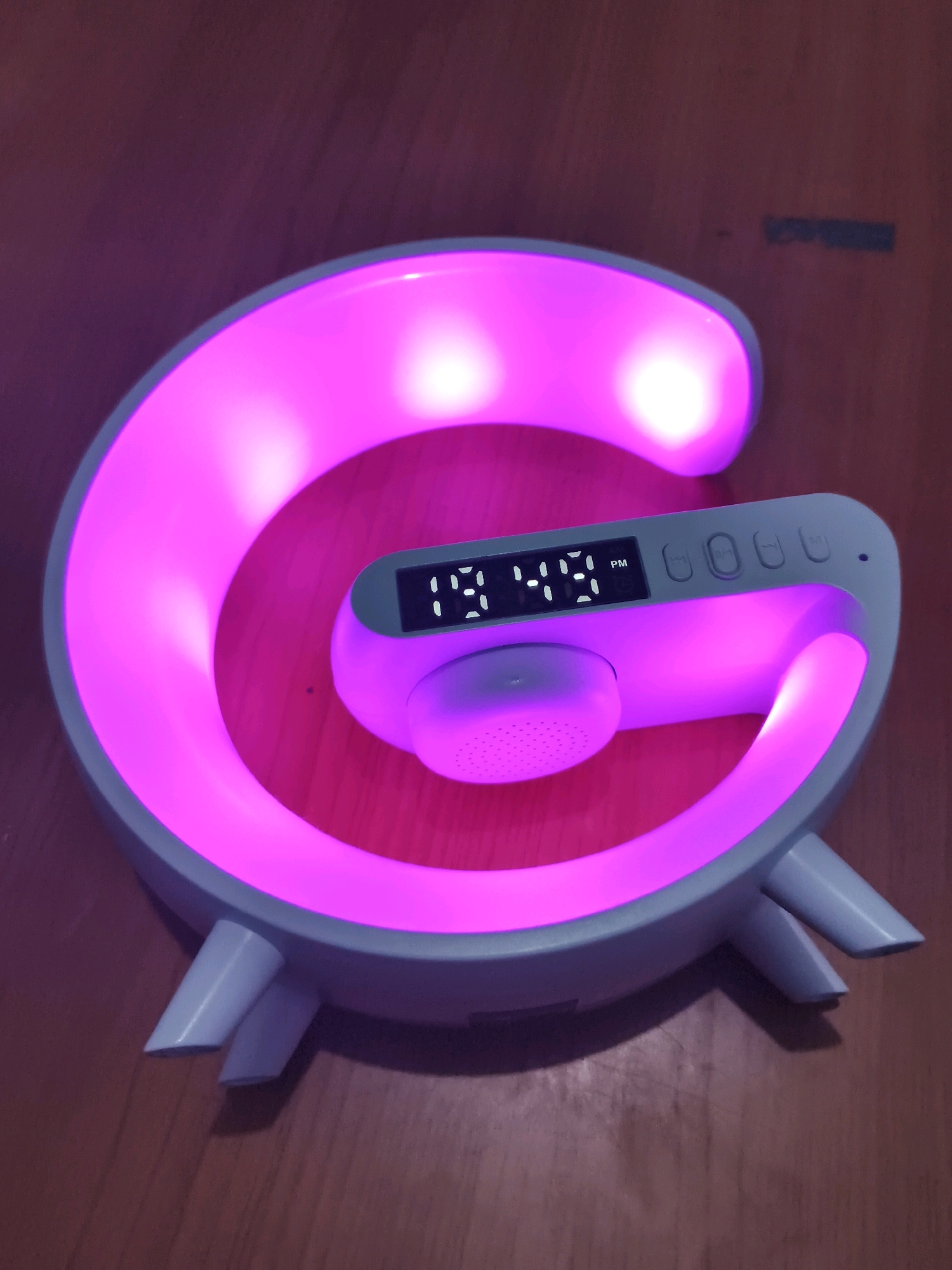 A pink glow emanates from a "G"-shaped lamp with a digital clock displaying "19:49 PM". The lamp has several buttons along its top and a speaker on its front. The lamp is resting on a wooden surface. The image appears to be taken indoors, in Bulakelor, Indonesia.