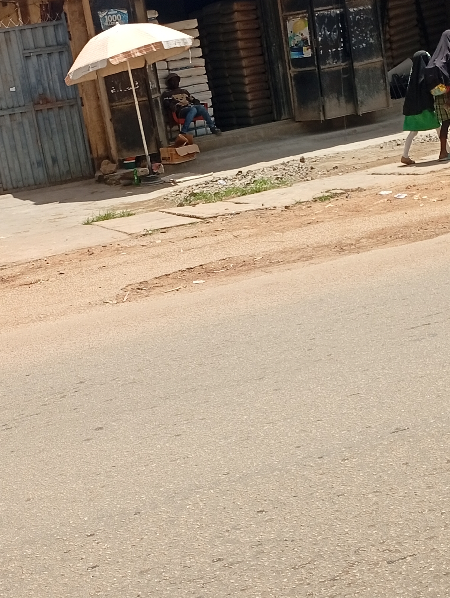 A man sits on a red chair under a beige and brown umbrella outside a building in Jos, Nigeria. The man wears a dark shirt with a graphic print and blue jeans. To his left, a brown cardboard box rests on the ground, and to his right, a green plastic bottle and a red container are visible. The building behind him has a metal door and appears to be a shop. A stack of what looks like cement bags is visible to the right of the building entrance. A young girl in a green skirt and a dark headscarf walks past on the sidewalk in front of the shop, accompanied by another person partially visible. The street in front of the sidewalk is paved with asphalt and shows some wear and tear. The overall scene depicts a typical street view in a Nigerian city.