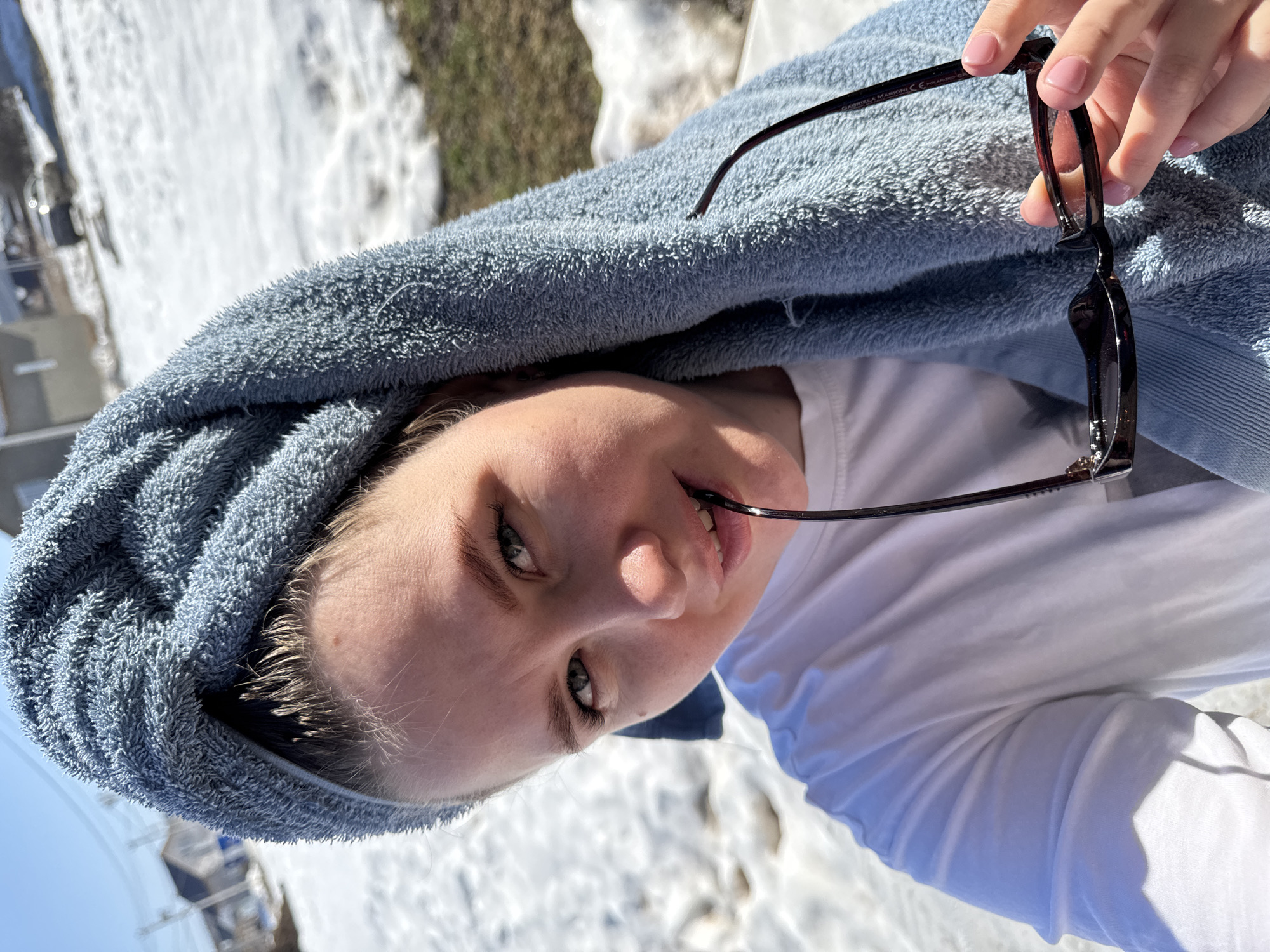 A selfie depicts a fair-skinned individual with light hair, draped in a light blue towel, holding black-framed eyeglasses in their right hand, and a portion of the eyeglass frame is held in their mouth. The person is wearing a white shirt. The background shows a snow-covered landscape, possibly in the vicinity of Gornyi (Dvurechenskoe S/P), Russia, with visible buildings and bare trees in the distance. The sunlight indicates it is likely daytime.
