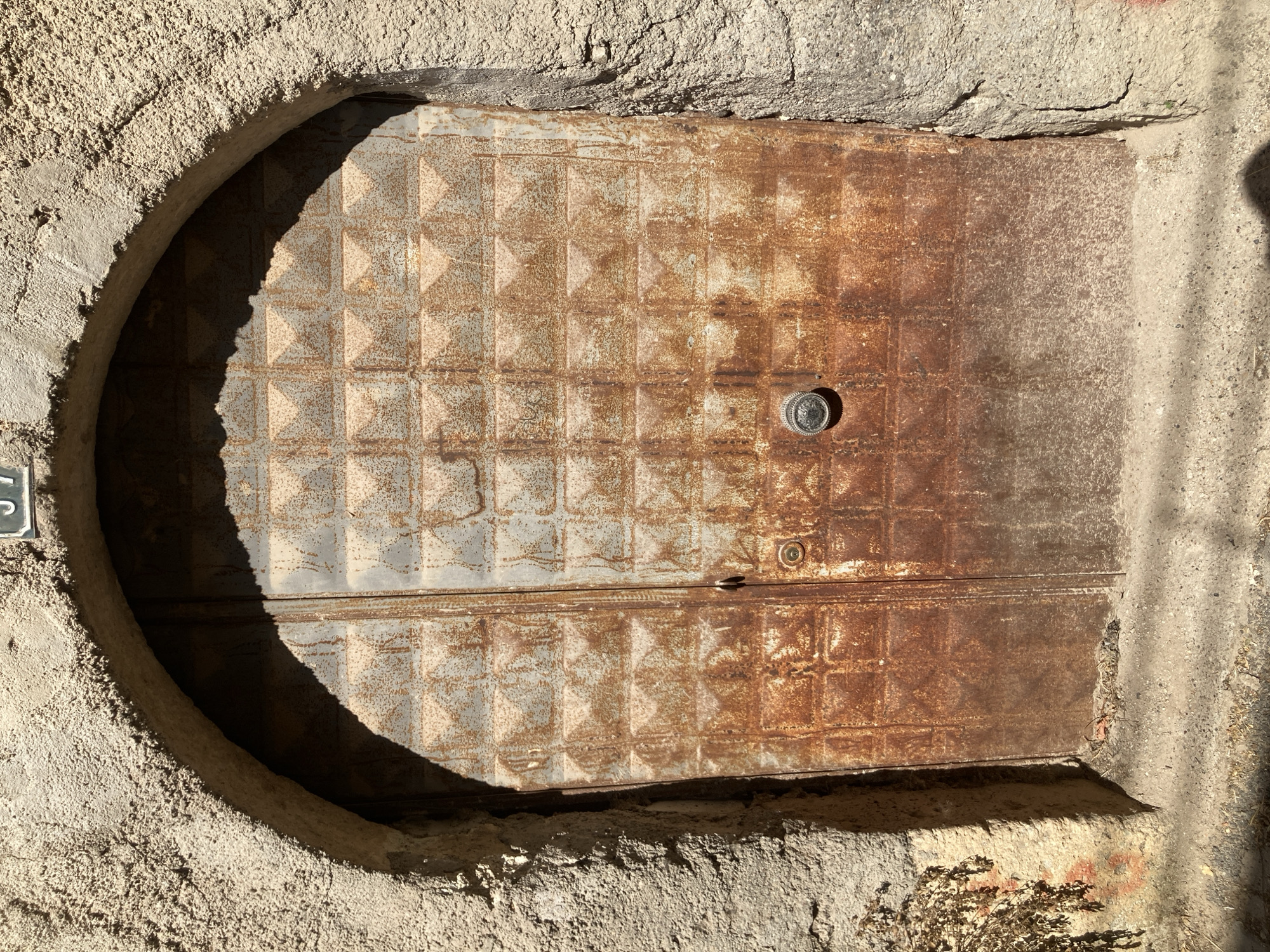 An aged, weathered arched doorway with a heavily rusted metal double door stands against a textured, rough-hewn stone wall in Avanos, Turkey. The door is segmented into a grid of square panels, each with a raised diamond pattern. A round, ornate metal doorknob is centrally located on the right panel, with a smaller, circular element above it. Sunlight casts a strong shadow across the upper portion of the door and the interior of the archway. The surrounding stonework is coarse and uneven, with a few dried plant remnants clinging to the lower left side. A small house number, "39", is affixed to the wall above the archway. The ground in front of the door is a mixture of gravel and dirt.