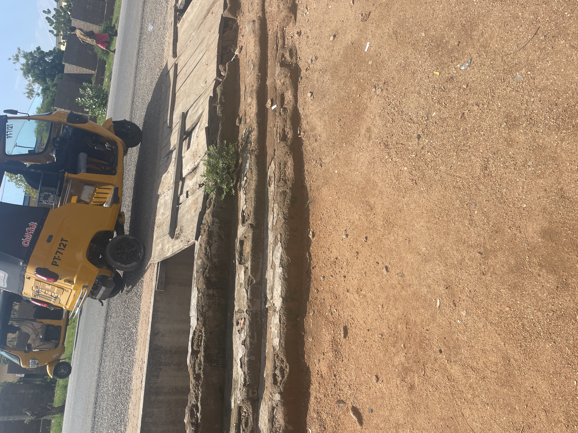 The image depicts an outdoor scene, likely in Jalingo, Nigeria, characterized by a dusty, reddish-brown terrain in the foreground. A dilapidated concrete structure with visible rebar and crumbling edges runs horizontally across the midground. A wooden plank bridge or cover spans part of this structure. In the background, two yellow auto-rickshaws (Keke Napep) are visible on a paved road. One auto-rickshaw is closer to the camera and features the license plate "PT-712T" and the word "Chikadi" in script. A person is seated in the driver's position of this auto-rickshaw. The second auto-rickshaw, further down the road, has a driver also seated. A pedestrian, wearing a checkered shirt and pink shorts, walks away from the auto-rickshaws on the right side of the road. The environment includes sparse vegetation and buildings in the distance. The sky is bright and clear, suggesting daylight.