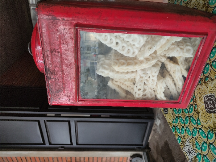 The image captures a close-up, rotated view of a rustic food display in what appears to be Kepanjen, Indonesia. The main subject is a rectangular, weathered red display case with a clear glass front. Inside the case, a pile of white, porous, and irregularly shaped snacks, strongly resembling Indonesian krupuk (crackers), is visible. The red paint on the display case shows signs of wear, with chipping and dark discoloration, suggesting age or frequent use.

To the right of the display case, a colorful patterned fabric is present, featuring traditional Indonesian batik motifs with floral and geometric designs in shades of yellow, green, and brown. Below the display case, a dark-colored piece of furniture, possibly a cabinet or counter, is visible, characterized by several clean, rectangular panels. At the very bottom edge of the frame, a segment of a reddish-brown brick wall can be seen.

No people or specific activities are depicted, but the setup implies a food vending or serving environment. The glass of the display case has some smudges, adding to the authentic, lived-in feel of the scene. There is no visible text in the image. The overall impression is one of a local, unpretentious food establishment.