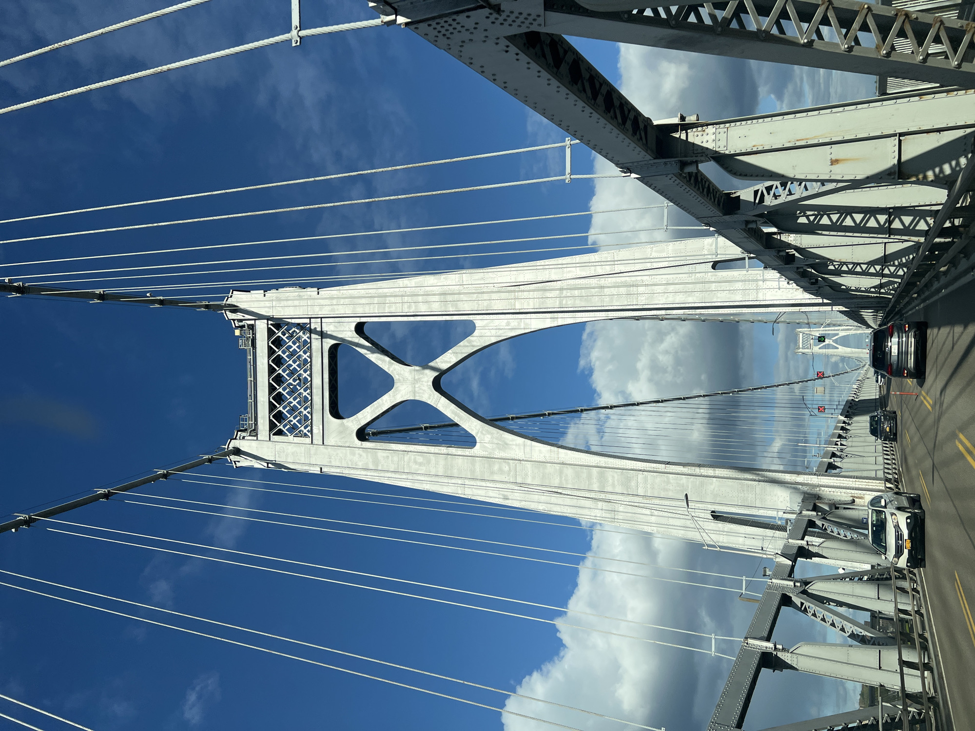 This image captures a vibrant daytime view from a vehicle traveling across a large suspension bridge. The perspective is from the road level, looking up and forward, showcasing the bridge's impressive steel structure against the sky.
The primary subjects are the bridge itself, the sky, and several vehicles. The bridge is constructed of robust grey steel girders, interconnected by rivets and intricate lattice work. Numerous white suspension cables run diagonally from the main towers and top structure down to the bridge deck, supporting the roadway. One of the main bridge towers is prominently featured on the left, displaying large, X-shaped or arched openings in its design.
The sky is a mix of deep blue and scattered white and grey clouds, indicating a partly cloudy day. Bright sunlight illuminates parts of the bridge and clouds, while other sections are in shadow, highlighting the three-dimensional complexity of the structure.
On the bridge deck, several cars are visible, including a white SUV and a dark sedan, suggesting active traffic flow. In the distance, faint red 'X' signs and a green light are discernible, possibly indicating lane closures or traffic signals.
Based on the provided location context, this is likely the Mid-Hudson Bridge in the City of Poughkeepsie, United States of America. The time of day appears to be mid-day to afternoon due to the clear lighting and shadows. No discernible text is clearly visible on any signs or objects within the frame. The overall scene conveys a clear, fair-weather journey across a significant piece of infrastructure.