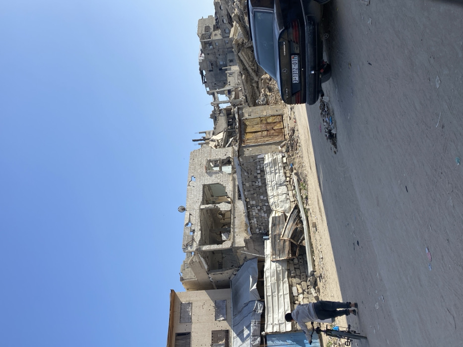 The image depicts a devastated urban street scene in Khan Yunis, Palestinian Territory, under a clear blue sky. In the foreground, an unpaved road with scattered debris and refuse recedes into the background. On the left, a lone individual, seen from behind, walks leftward, pushing a dark-colored bicycle. This person wears a light-colored hooded top and dark trousers.
To the right of the person and bicycle, a black Mercedes-Benz sedan (E-Class W124 model) is parked facing away from the camera. Its rear license plate reads "3-1312-01".
The background is dominated by severely damaged and partially collapsed multi-story buildings. On the immediate left, a light-colored building shows numerous small circular impact marks on its facade, with windows either boarded or covered. Next to it and extending into the mid-ground are structures with exposed interiors, broken concrete, and twisted rebar, indicative of significant structural damage or destruction. One partially standing structure prominently features a small, white satellite dish. A segment of a wall constructed from concrete blocks topped with corrugated metal fencing is visible, incorporating a rusted, double-leaf metal door. Further into the background, a dense collection of buildings is reduced to rubble and skeletal frameworks, confirming widespread destruction. The scene is illuminated by strong sunlight, casting distinct shadows.
