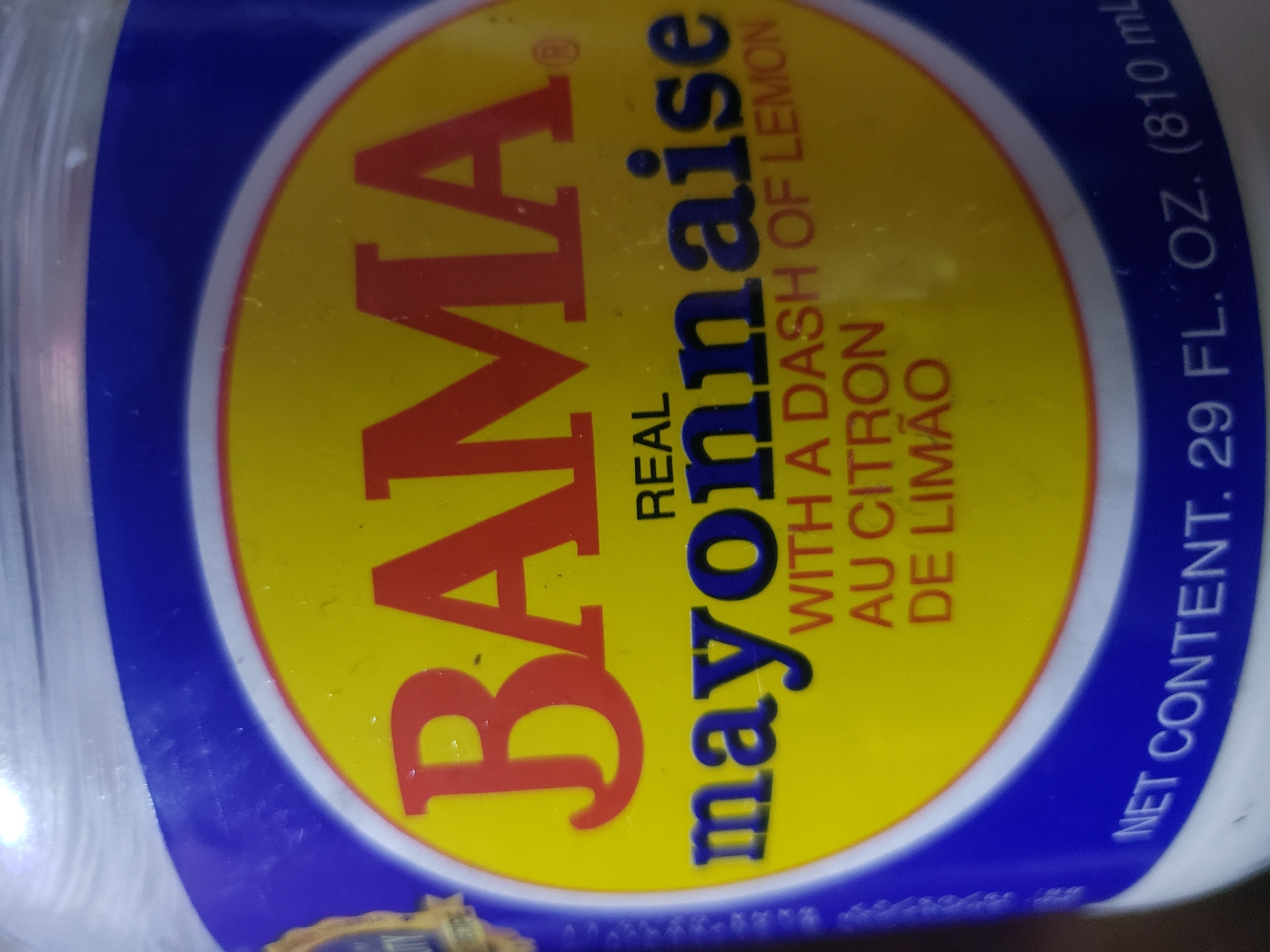 The image shows a close-up of a jar of BAMA Real Mayonnaise. The label is predominantly yellow and blue. Large red letters spell out "BAMA" at the top of the yellow section. Below this, in dark blue letters, is the word "mayonnaise". Smaller red text below that reads "WITH A DASH OF LEMON AU CITRON DE LIMÃO". Along the bottom of the label, in white text against a blue background, is "NET CONTENT. 29 FL. OZ. (810 ml)". The background is blurry and appears to be indoors, with some reflected light on the jar. There is no visible indication of Iganmu, Nigeria, or any specific city location.