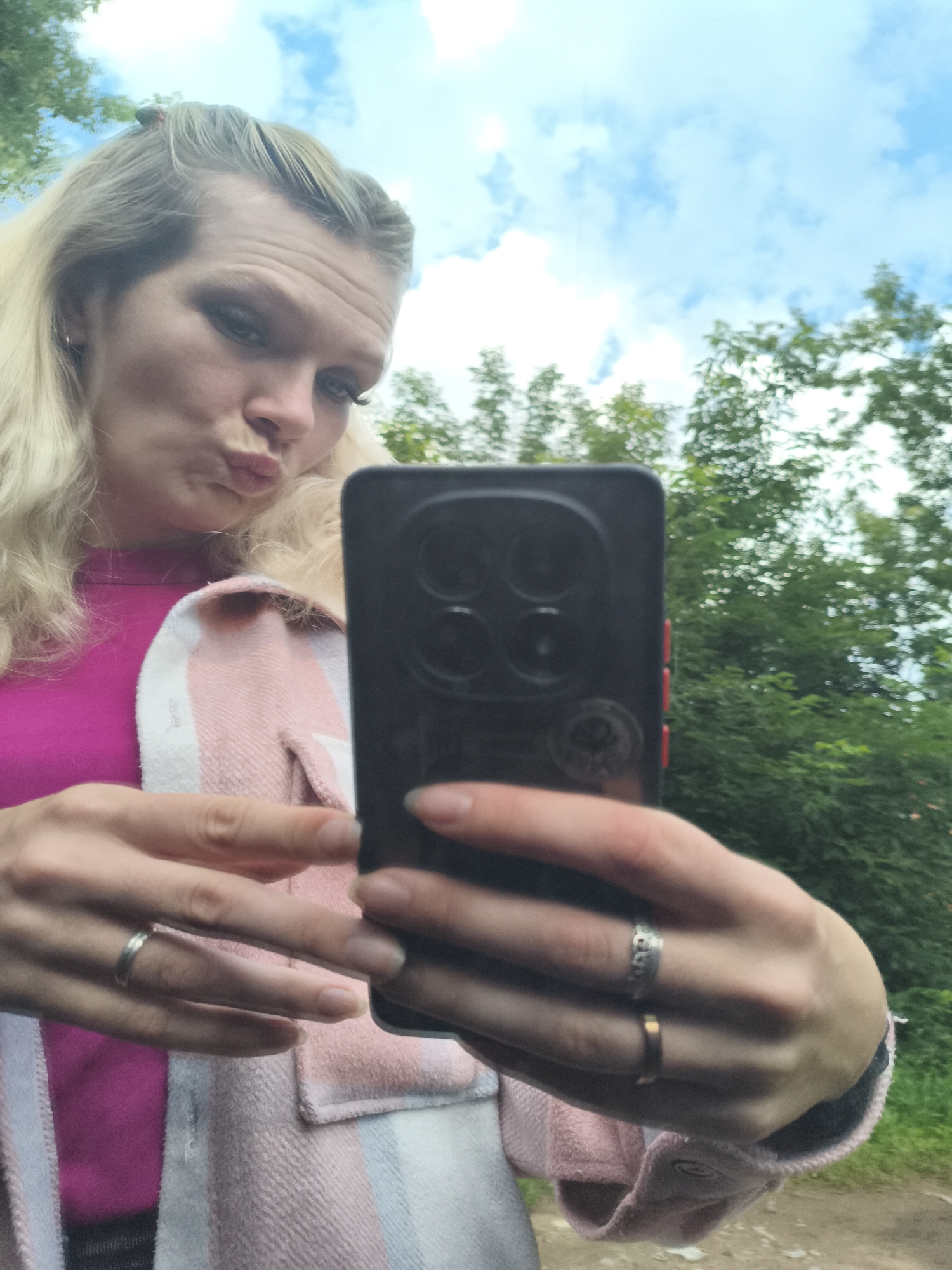 A young woman with blonde hair is taking a selfie outdoors. She is wearing a bright pink shirt and a pink and white checkered jacket. She is holding a black smartphone in her right hand, and her left hand is resting on her chin. Her lips are pursed, as if she is about to kiss the camera. The background is a blur of green trees and blue sky with white clouds. The location is not specified as Izhevsk, Russia.