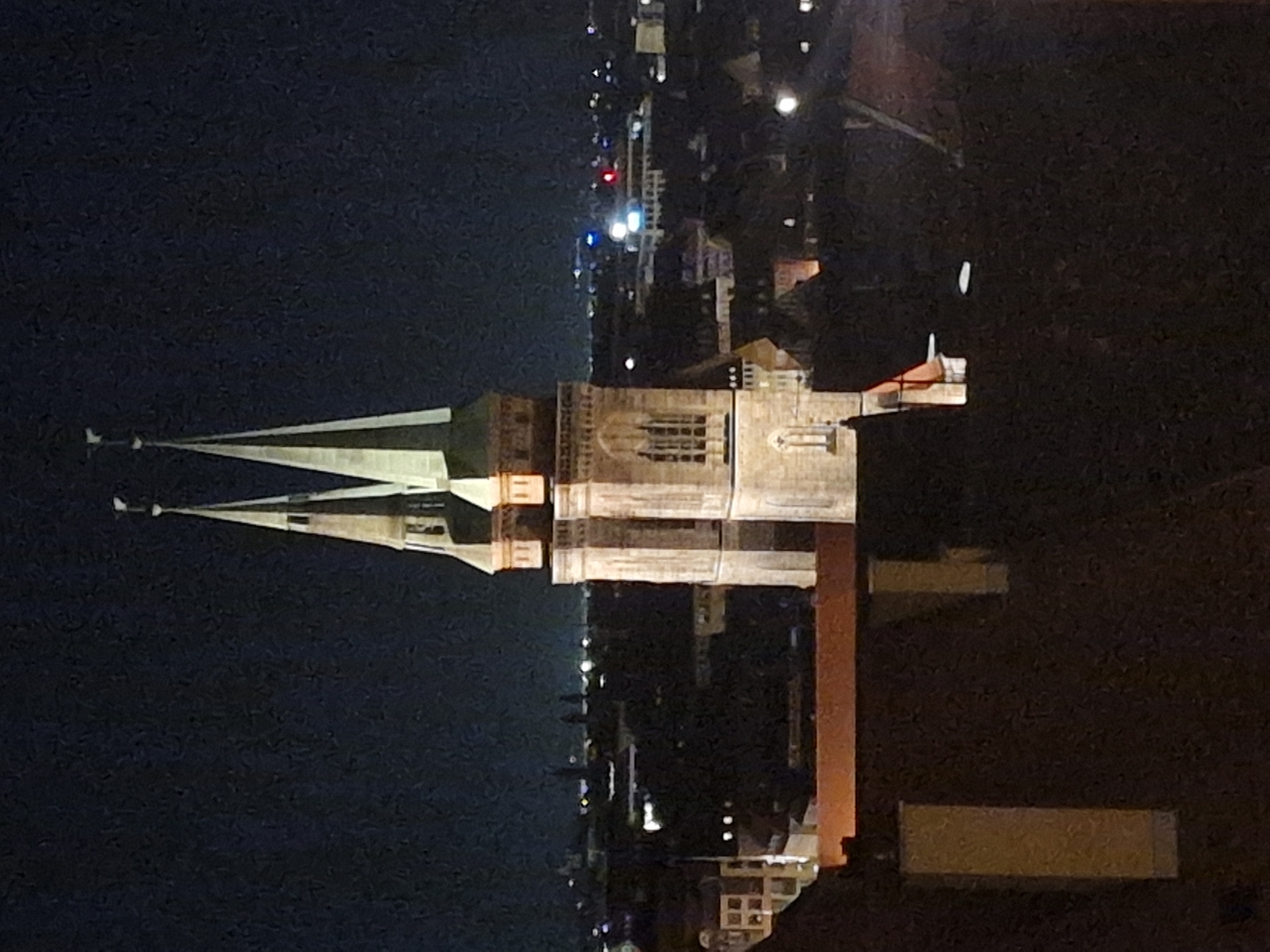 The image shows a night view of the Nuremberg skyline, with the iconic twin spires of St. Lorenz Church prominently featured in the foreground. The church's spires, illuminated by artificial light, rise against a dark night sky. Below the spires, the intricate stone facade of the church is visible, with arched windows and architectural details. In the background, the urban landscape of Nuremberg unfolds, with numerous buildings and lights dotting the horizon. The lower portion of the image reveals rooftops and structures in the immediate vicinity of the church, with warm-toned lights suggesting illuminated interiors. The overall atmosphere is one of a serene and majestic cityscape at night.