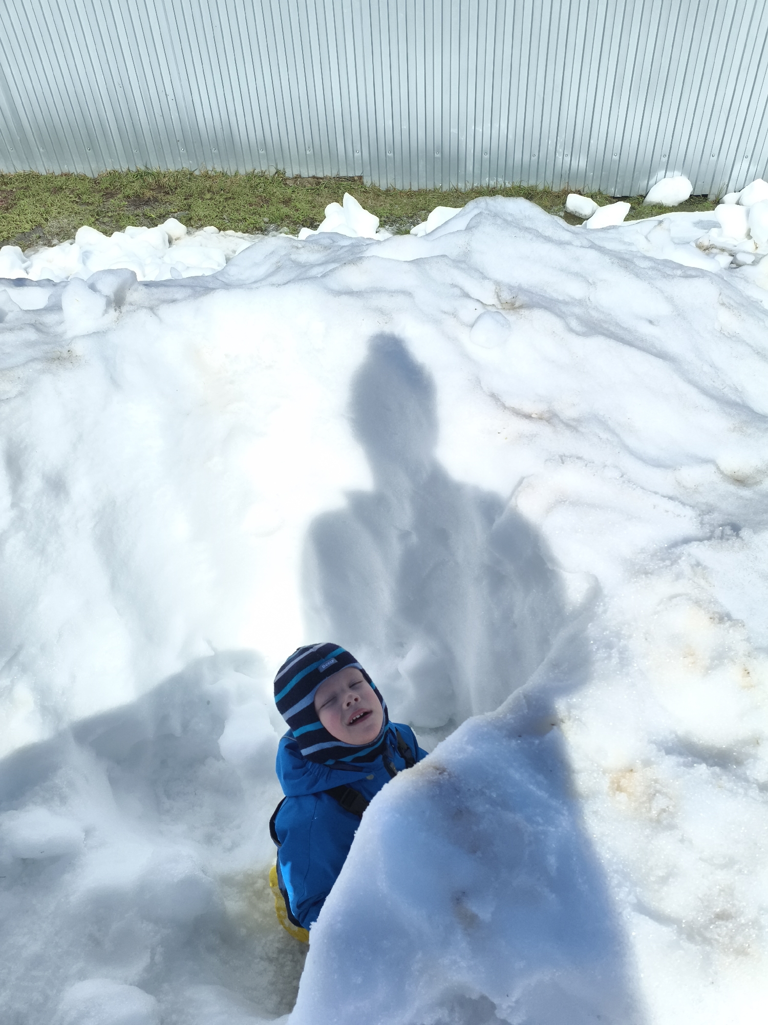 This vertical shot captures a young child playing in large snowdrifts on a bright, sunny day in Русский Вожой, Russia. The child, likely a boy, is the primary subject, positioned in the lower-middle of the frame, appearing to be standing or sitting within a significant accumulation of snow. He wears a blue winter jacket and a striped blue and black winter hat, which has the word "Beezy" visible on it. His head is tilted upwards and slightly back, with his eyes mostly closed and mouth slightly open, suggesting he might be looking towards the bright sun or enjoying the moment.
The scene is dominated by white snow, forming undulating mounds and drifts that fill most of the foreground and midground. Behind the child and across the snow, a large, distinct shadow of an adult (presumably the photographer) is cast, indicating a strong overhead light source. In the background, a light-colored, corrugated metal fence or wall spans the width of the image. Below the fence, a strip of vibrant green grass is visible, peeking out from beneath the melting snow, suggesting that it is either late winter or early spring. The bright sunlight and deep shadows highlight the texture of the snow and create a clear, crisp atmosphere.