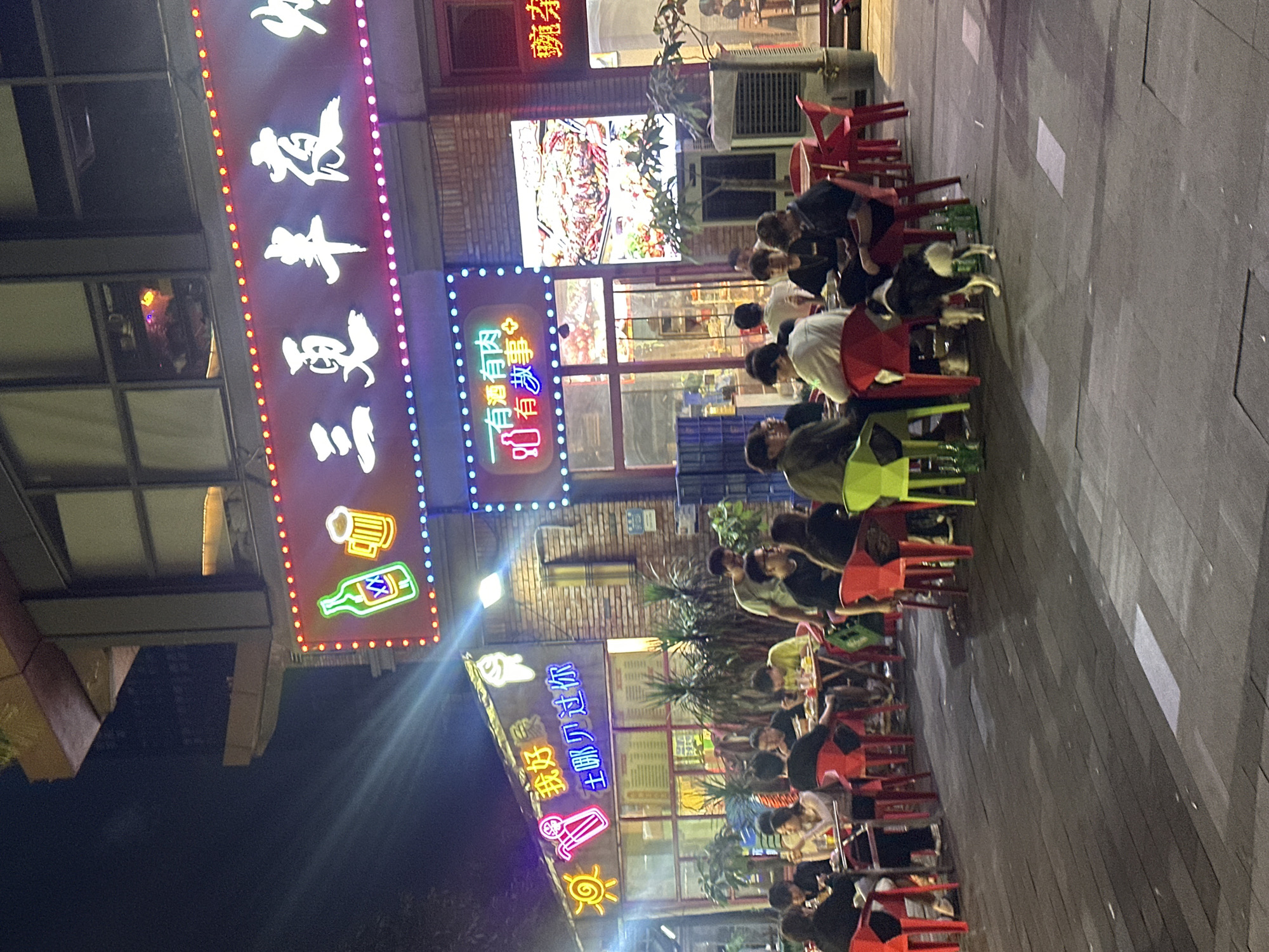 The image depicts an outdoor dining area in the Chongqing High-tech Zone, China, at night. Neon signs with Chinese characters illuminate the scene. Several groups of people are seated at tables, some eating and others socializing. A black and white dog is visible near one of the tables. The ground appears to be paved, and there are potted plants and an air conditioning unit in the background. The overall atmosphere suggests a casual dining establishment.