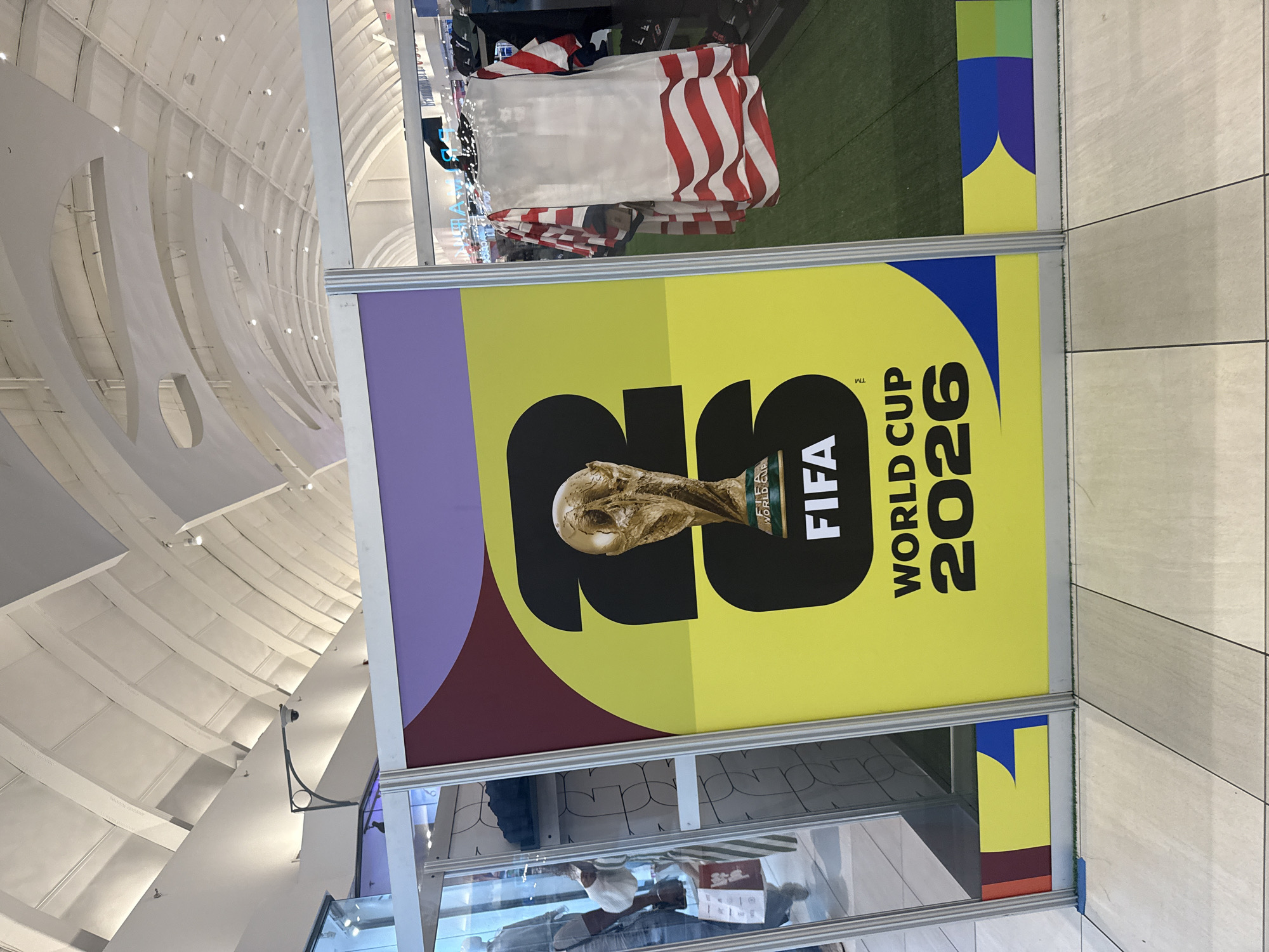 This image captures a promotional display for the FIFA World Cup 2026 situated within a large, brightly lit commercial interior, likely a shopping mall or airport terminal, in Sunrise, United States of America.
The central subject is a vertical display panel, framed in silver metal, showcasing the FIFA World Cup 2026 logo. The panel features a vibrant yellow background with a large black "26" graphic. Superimposed on the "6" is an image of the golden FIFA World Cup trophy. Below the trophy, the word "FIFA" is printed in white letters on a black rectangle, followed by "WORLD CUP 2026" in black text. The top section of the panel is purple, and a maroon band separates it from the yellow. The bottom border of the panel contains abstract colorful shapes in maroon, yellow, and blue.
To the immediate right of the display, a retail section is visible, featuring clothing items hung on racks. A prominent white sleeveless garment with red wavy stripes, resembling a patriotic or flag design, is displayed. The floor in this retail area is covered with green artificial turf. Further into the background, additional clothing and display units are visible.
To the immediate left of the display, a glass panel reflects part of the environment. A person wearing a light-colored shirt and blue jeans, carrying a red shopping bag, is reflected walking away from the viewer. Another partial reflection shows a person wearing a white hat or head covering. The floor in the foreground consists of light-colored square tiles with dark grout lines.
The expansive ceiling above is vaulted and white, constructed with a series of parallel, arched structural elements and visible cross-beams, characteristic of a large public space. Numerous small, circular recessed lights illuminate the area. A white dome-shaped security camera is mounted on a black, curved bracket extending from a wall segment in the upper left portion of the image. The overall lighting is bright and even.