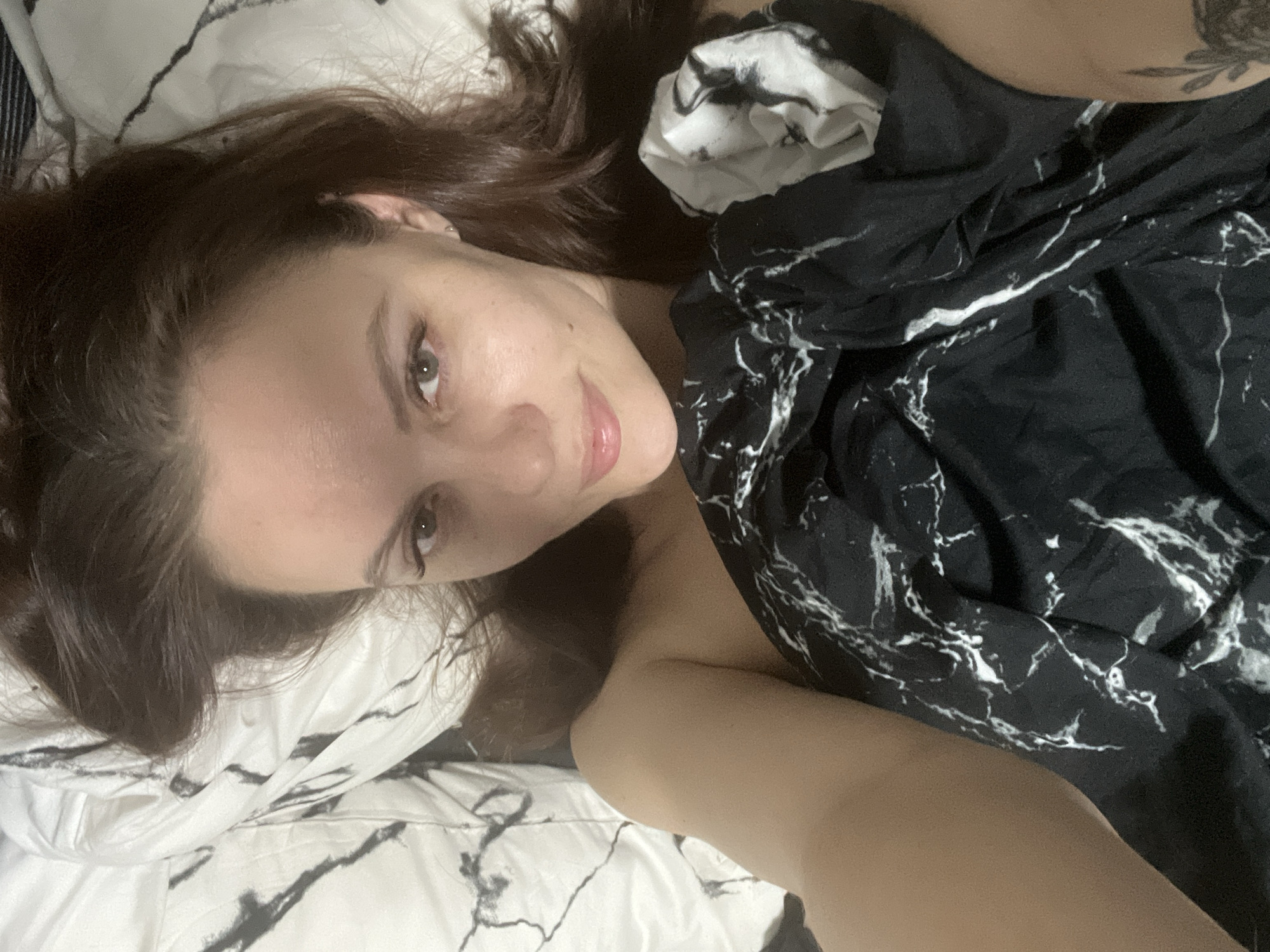 A young woman with long, dark brown hair is captured in a close-up, angled shot, lying in bed. She looks directly at the camera with light-colored eyes (appearing greenish-grey) and a slight, gentle smile. Her lips are soft and appear glossed. A small mole is visible on her right cheek, near her nose. She appears bare-shouldered or partially covered, resting her head on a white pillow with a black, marble-like pattern.
Her body is partially obscured by a black duvet or blanket featuring a prominent white marble pattern. A tattoo with a floral or leafy design is subtly visible on her right upper arm, near the top right of the frame. The lighting is soft and dim, suggesting an indoor setting, most likely a bedroom, possibly in the evening or early morning. The overall atmosphere conveyed is relaxed and intimate. The scene is situated in Chaikovsky, Russia. There is no visible text in the image.
