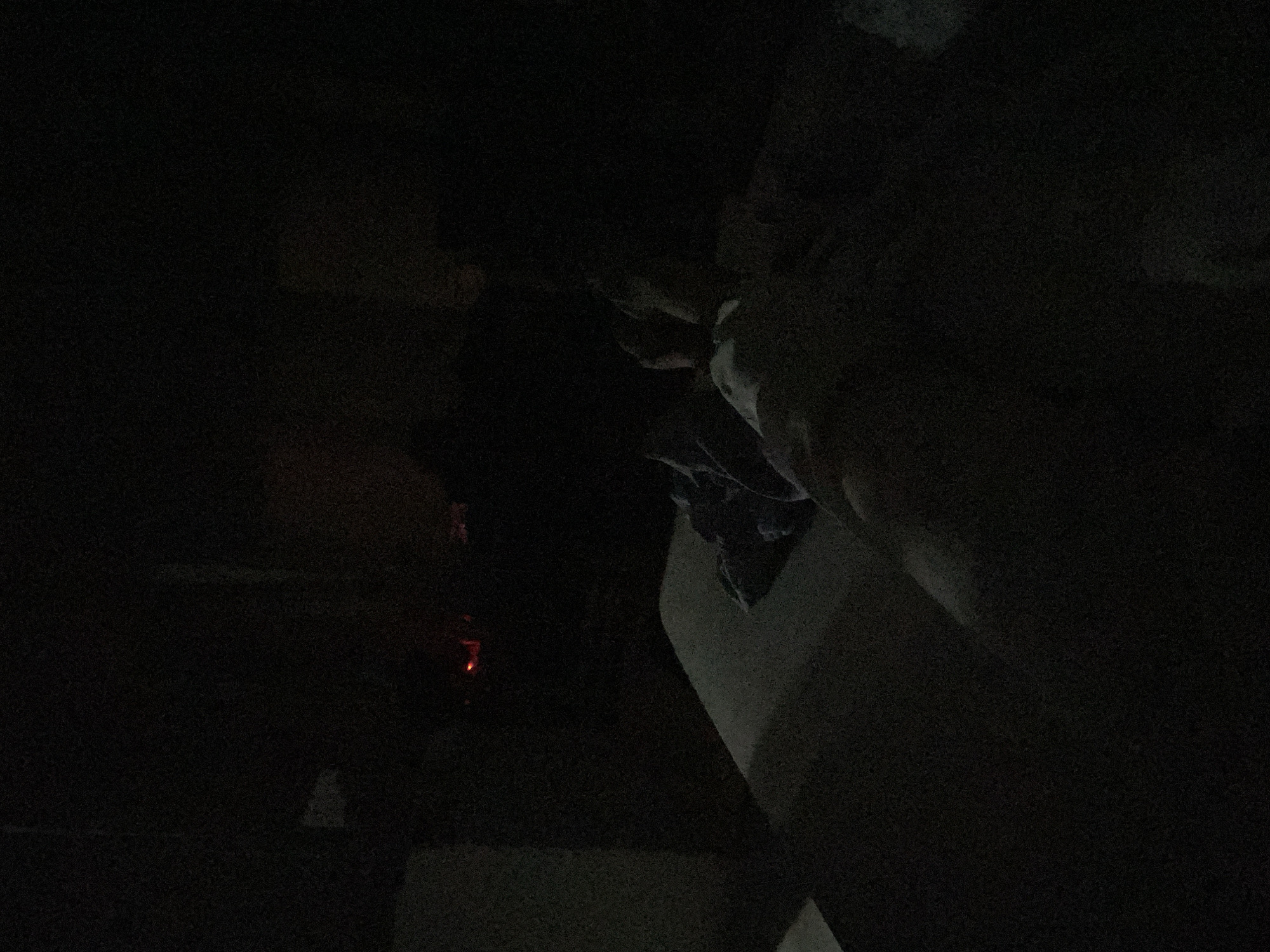 I am unable to determine the location of this image.
The image appears to be taken in a dark, indoor environment. A faint, red light is visible towards the left. A pale object with what appears to be fabric on it is present on the right.