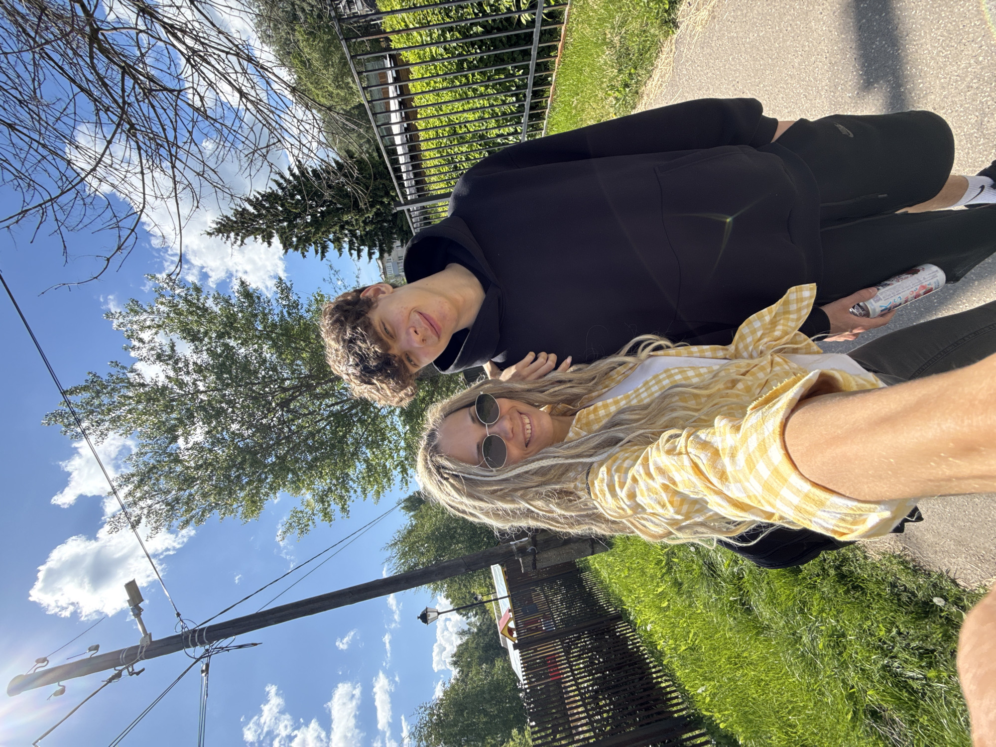 The image captures a bright outdoor selfie featuring two young individuals, a man and a woman, on a sunny day in Rusanovo, Russia. The photo is taken from a low angle, looking upwards towards the clear blue sky dotted with white cumulus clouds.
On the left, a young man with dark, curly hair wears a black hoodie and black sweatpants, which feature a white Nike swoosh logo on the left leg. He holds a white beverage can adorned with faint red, white, and blue graphics and text that appears to include "JAPANESE STYLE." He offers a subtle smile to the camera.
Beside him, a young woman with long blonde hair, styled in dreadlocks or braids, is seen wearing round sunglasses and a cheerful yellow and white checkered long-sleeved shirt. She smiles broadly at the camera.
The background reveals a vibrant green environment with various trees, including both deciduous and coniferous types. A black metal fence separates a grassy area from a paved path where the subjects are positioned. A prominent utility pole with wires and electrical equipment stands on the left, with the sun creating a bright lens flare near its base. A street lamp post is also visible further back behind the woman. The overall impression is one of warmth, relaxation, and happiness, suggesting a pleasant outdoor activity.