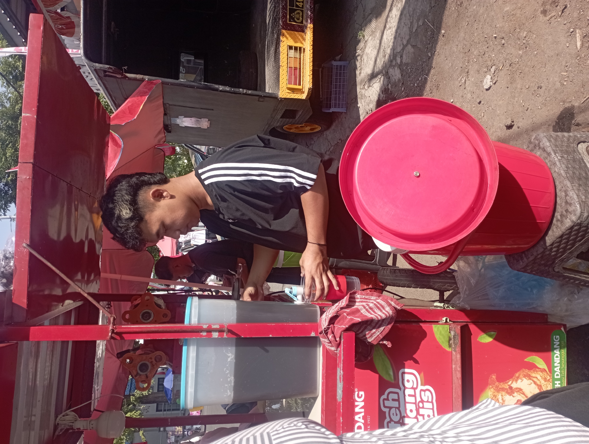 The image captures an outdoor scene in Nganjuk, Indonesia, likely a street or roadside. In the foreground, a young person with a distinctive hairstyle, wearing a black t-shirt with white stripes on the sleeves, is serving a beverage. They are holding a clear cup and appear to be filling it from a dispenser on a mobile cart.
The cart is predominantly red and features prominent branding that includes "DANG" and what appears to be "Teh Nganjuk" along with text in Indonesian. Two large, clear plastic containers filled with a dark liquid, presumably tea, are visible on the cart. A red fabric cloth with white stripes is draped over a part of the cart.
In the background, parts of a truck are visible, with yellow and black accents on its rear. The ground is paved with a rough, cracked surface, and some debris is scattered around. Trees and foliage are present in the middle ground, suggesting an urban or semi-urban environment. Sunlight casts shadows, indicating it is daytime. The overall impression is of a street vendor operation, likely serving refreshing drinks.