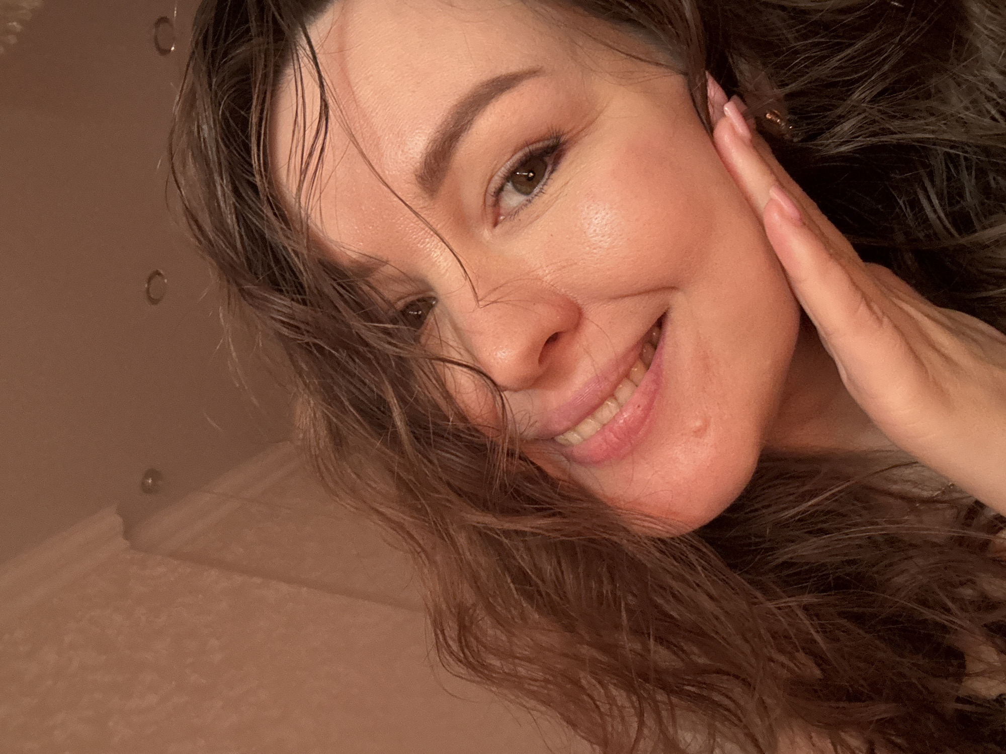 A close-up portrait shows a smiling woman with long, dark, and seemingly wet or damp wavy hair. She is angled towards the camera, looking directly at the viewer with a joyful and bright expression. Her left hand is gently placed on her cheek, near her ear, with manicured nails visible. Her skin appears fresh and radiant, and her eyes are bright. The background is a soft, uniformly light, warm-toned surface, possibly a wall or ceiling, featuring two discernible small, circular, recessed objects that could be lights or fixtures, hinting at an indoor setting such as a bathroom. The lighting is soft and warm, contributing to a pleasant and intimate atmosphere. The woman's demeanor conveys happiness and approachability. No text is visible in the image. The location context for this media file is Yuzhny, Russia.