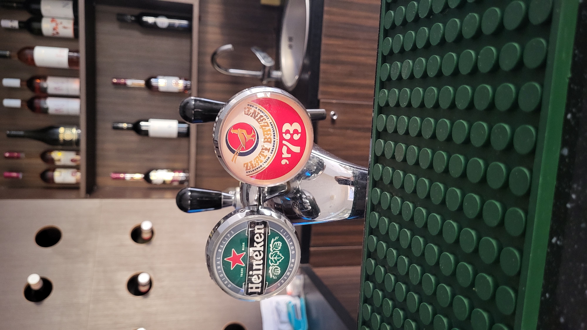 The image depicts a close-up of two beer taps in what appears to be a bar or establishment. The tap on the left features the Heineken logo, a green circular emblem with the brand name and a red star. The tap on the right displays a red circular emblem with a golden pheasant and the text "ZLATÝ BAŽANT '73". Both taps are made of polished metal and have black handles. In the background, shelves are visible, stocked with various bottles, likely wine and spirits. A sink and faucet are also present in the background. The foreground shows a textured green surface, possibly a drip mat. The overall scene suggests a commercial setting where alcoholic beverages are served. The image does not provide any specific visual clues to pinpoint its location within the city of Prešov, Slovakia.