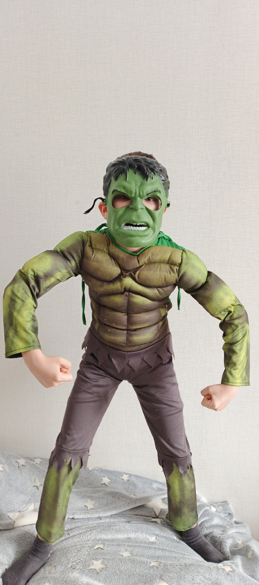 A child is depicted in an indoor, full-length portrait in Chaikovsky, Russia. The subject is wearing a Hulk-themed costume. The attire includes a rigid green mask with an angry facial expression, simulated black hair, and eye cut-outs revealing the child's eyes. The mask is secured by black elastic bands extending behind the ears. The upper body costume component is padded green and brown fabric, simulating a muscular physique across the chest and arms. A green fabric element is present around the neck. The lower body consists of dark grey trousers with ragged, jagged hems at the waist and mid-calf, worn over dark grey socks. The child is standing with legs slightly apart and both hands clenched into fists at hip level. The background features a plain, light-colored wall with a subtle texture. The child stands on a rumpled light grey blanket adorned with numerous white star patterns.
