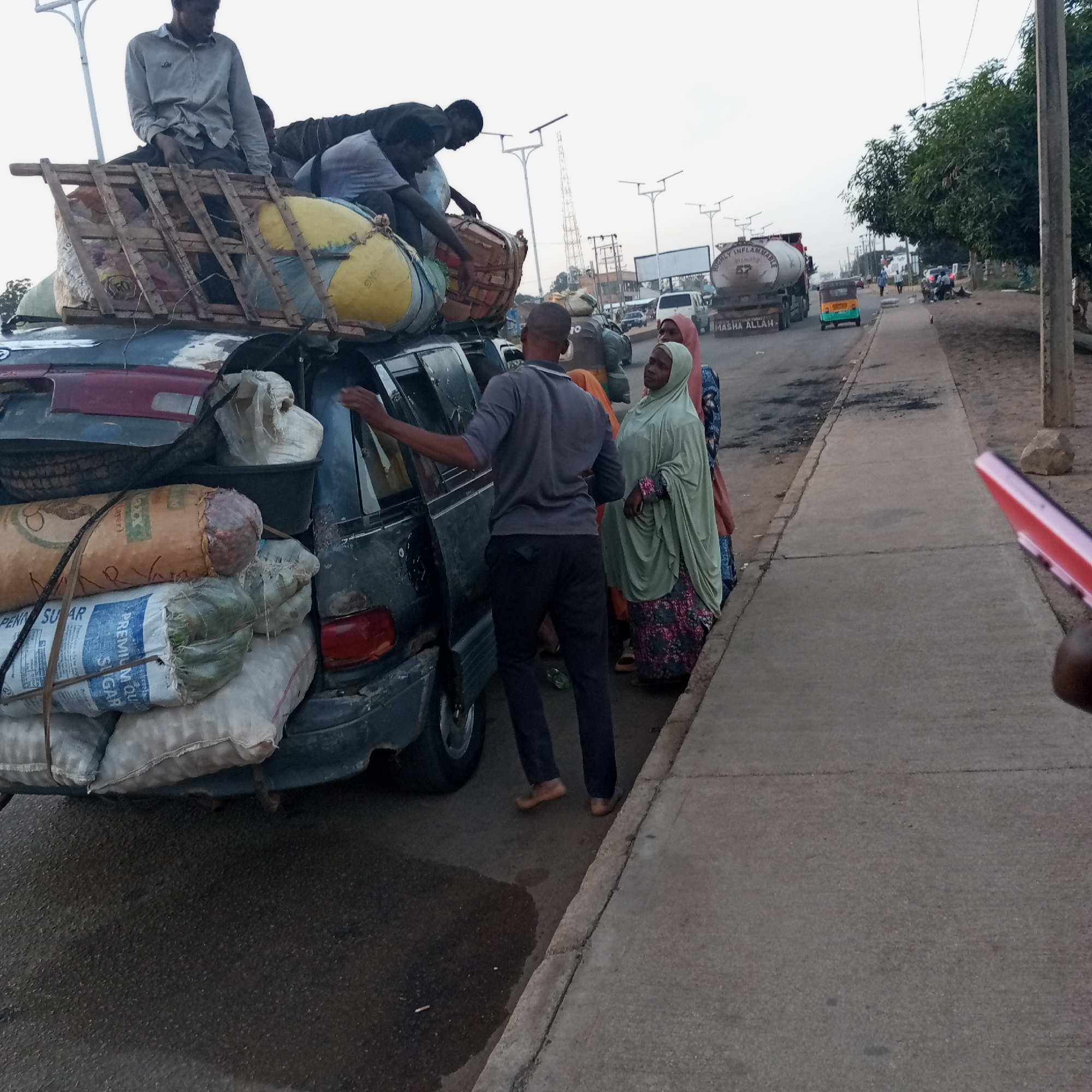 The image shows a dark-colored minivan severely overloaded with goods, including sacks labeled "PREMIUM SUGAR" and other miscellaneous items strapped to the roof with wooden framing, with several people on top of the load, on a street in Du, Nigeria. A man in a gray shirt and black pants stands by the open door of the van. Several women in hijabs and colorful patterned dresses stand on the sidewalk nearby. In the background, a tanker truck labeled "MASHA ALLAH" and "HIGHLY INFLAMMABLE" can be seen, along with other vehicles and buildings lining a wide street with streetlights. A sidewalk runs alongside the road where the minivan is parked.